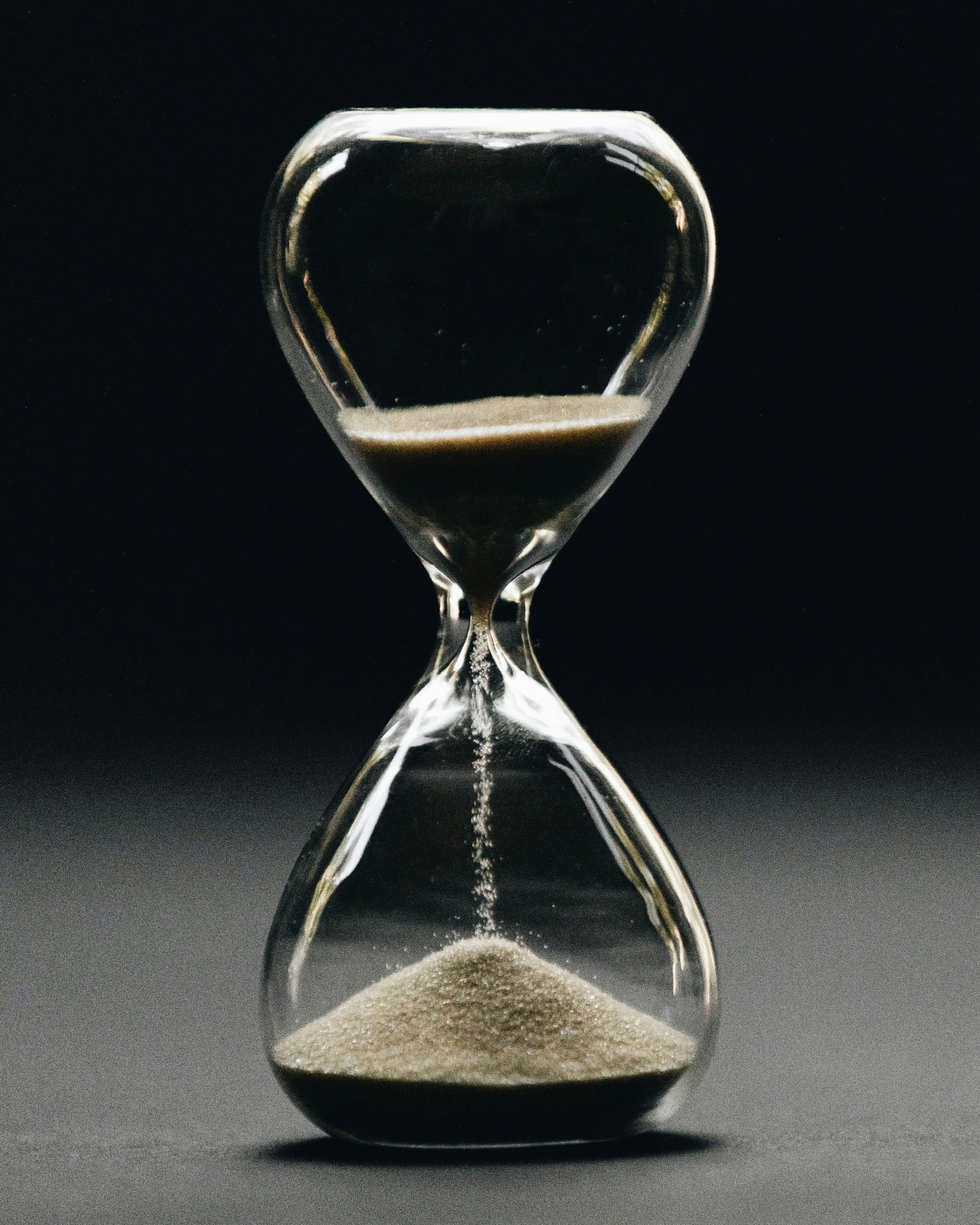 An hourglass with black sand, with some sand flowing from the top chamber to the bottom chamber against a dark background. This represents the element of recruitment and search which is interim recruitment. You are under time pressure.