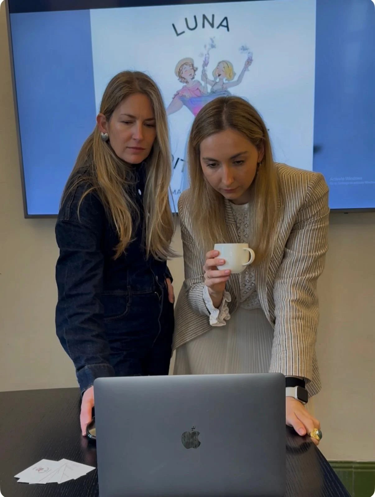 Two women are looking at a laptop on a table, one holding a mug, with a large screen behind them displaying a book cover titled 'LUNA' and an illustration of two girls blowing smoke.