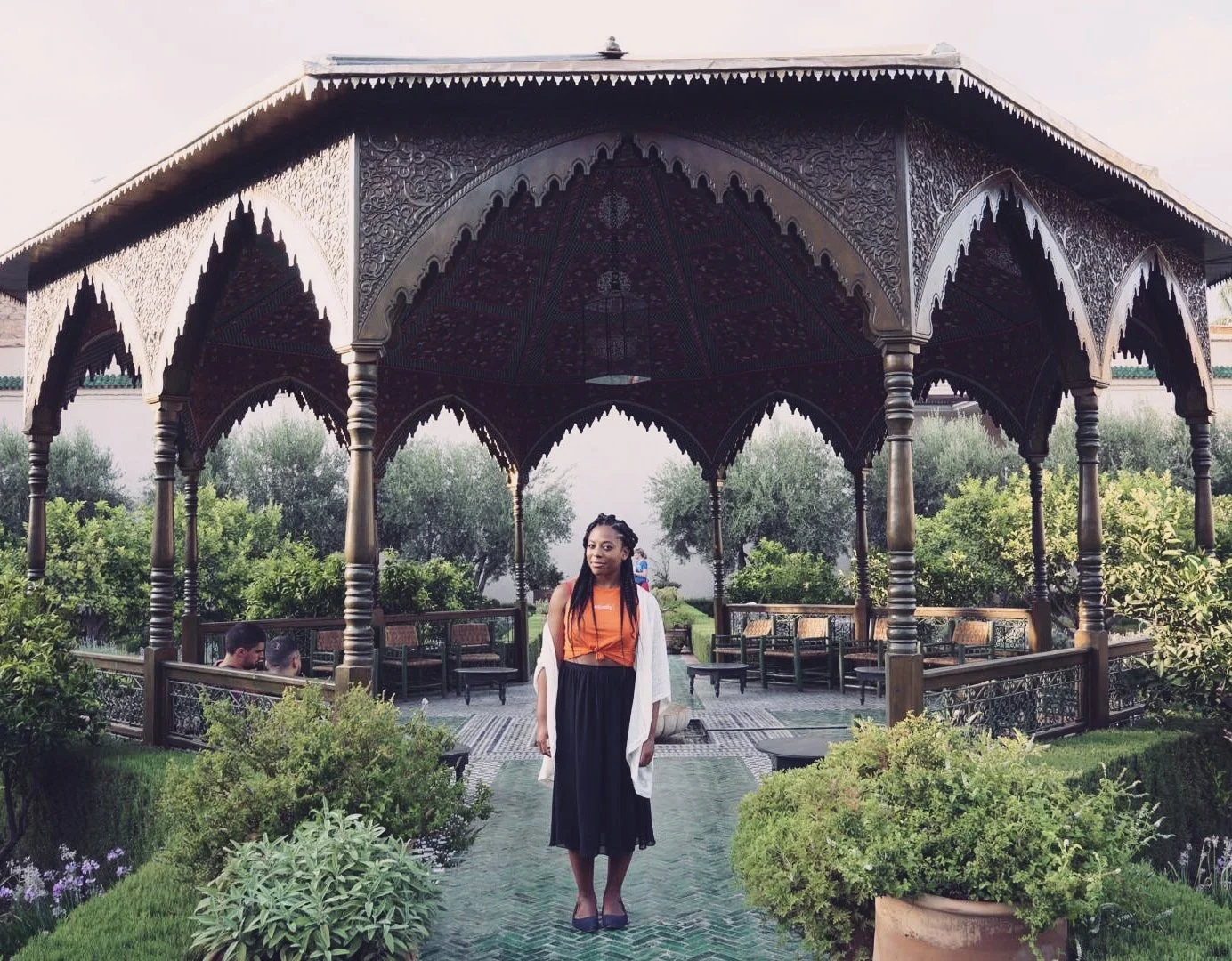 A woman with long braids wearing an orange top and black skirt standing under a decorative gazebo with lush greenery and trees in the background.