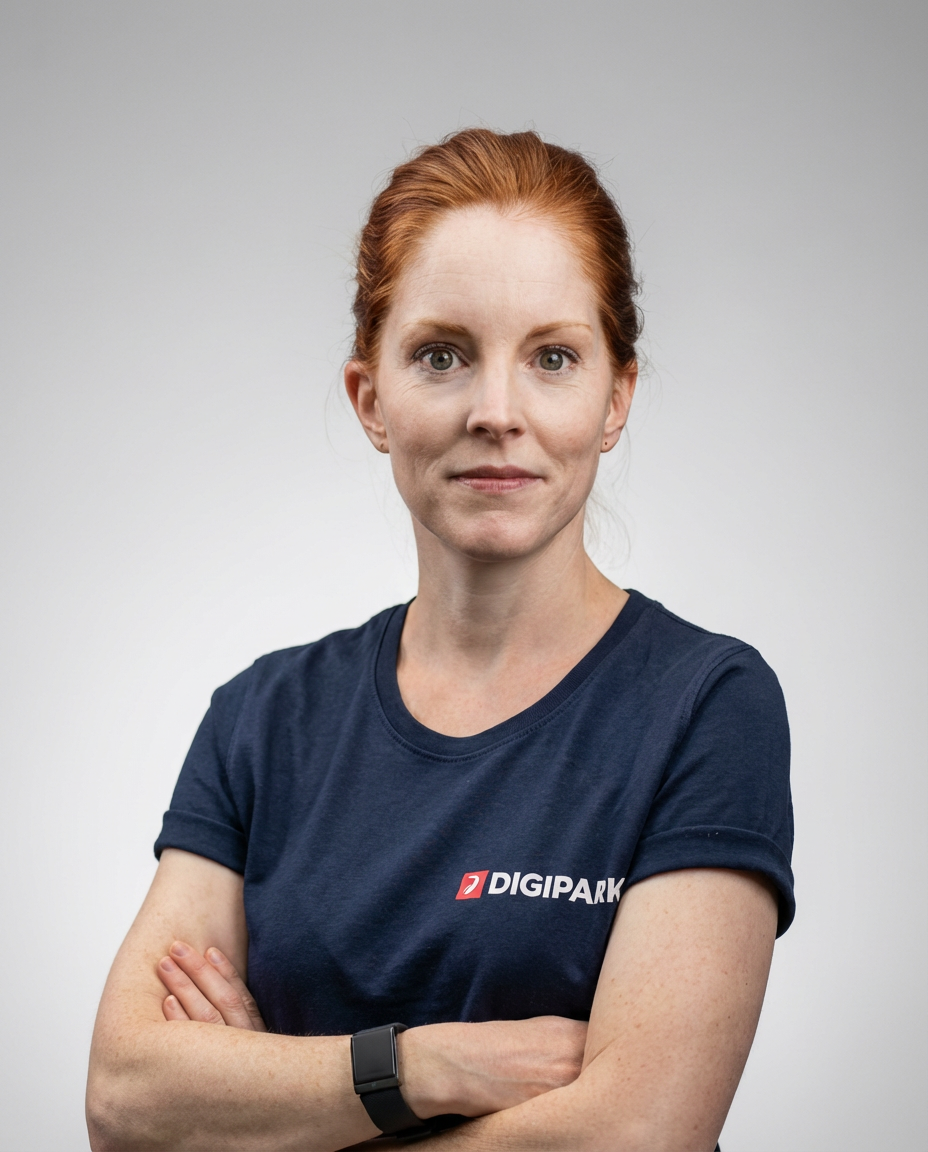Das Bild zeigt Maren Frankemölle, Head of Sales & Marketing. Eine Frau mit roten Haaren, die ein dunkelblaues T-Shirt trägt mit dem Logo 'DIGIPARK', steht vor einem grauen Hintergrund, mit verschränkten Armen und schaut direkt in die Kamera.