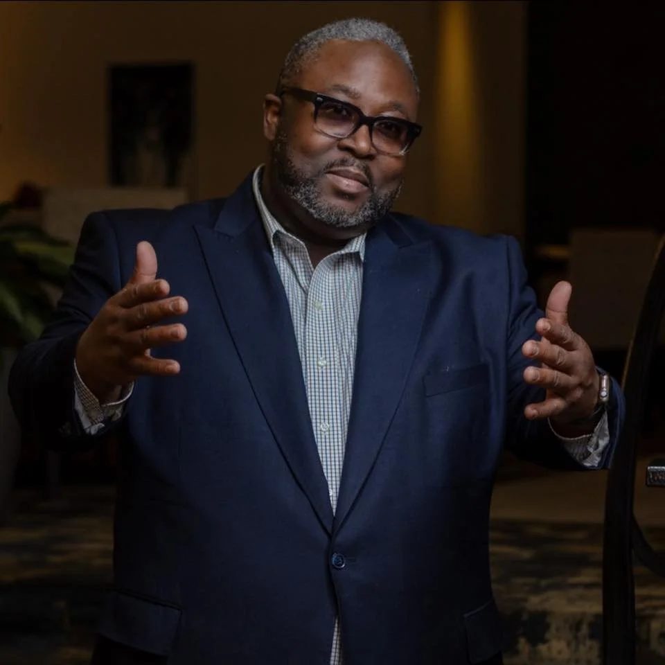 Man in a blue suit and glasses making gestures with his hands in a dimly lit conference or event setting.