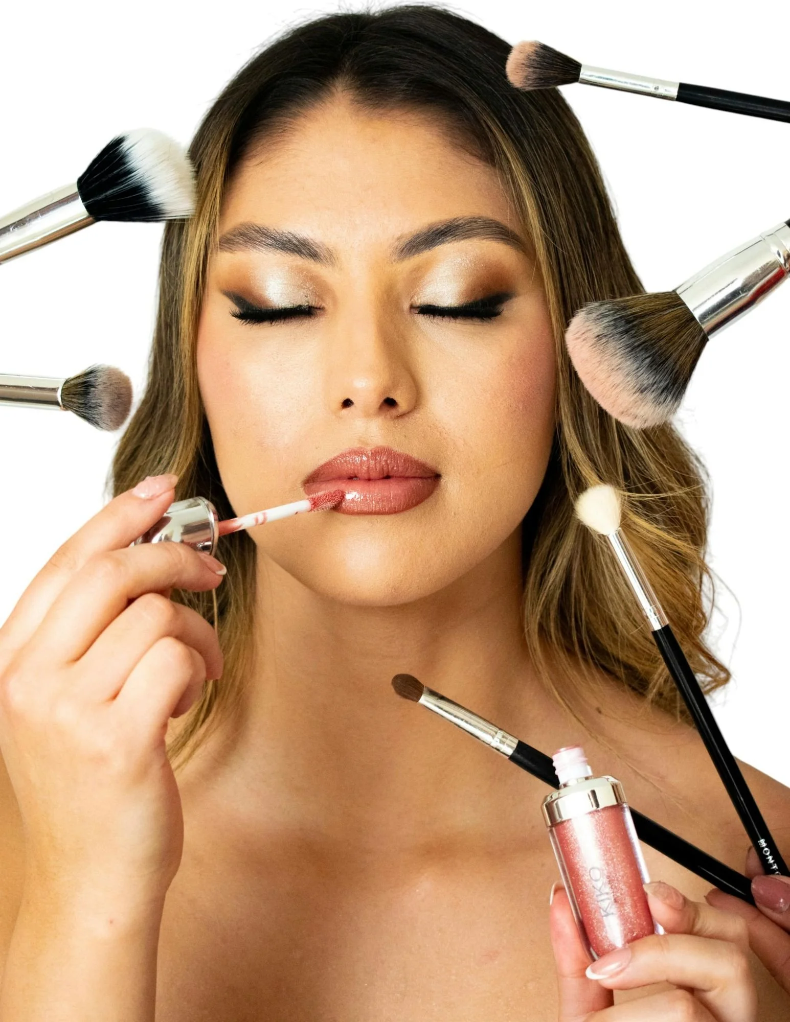 Woman with eyes closed, applying lip gloss while surrounded by makeup brushes.