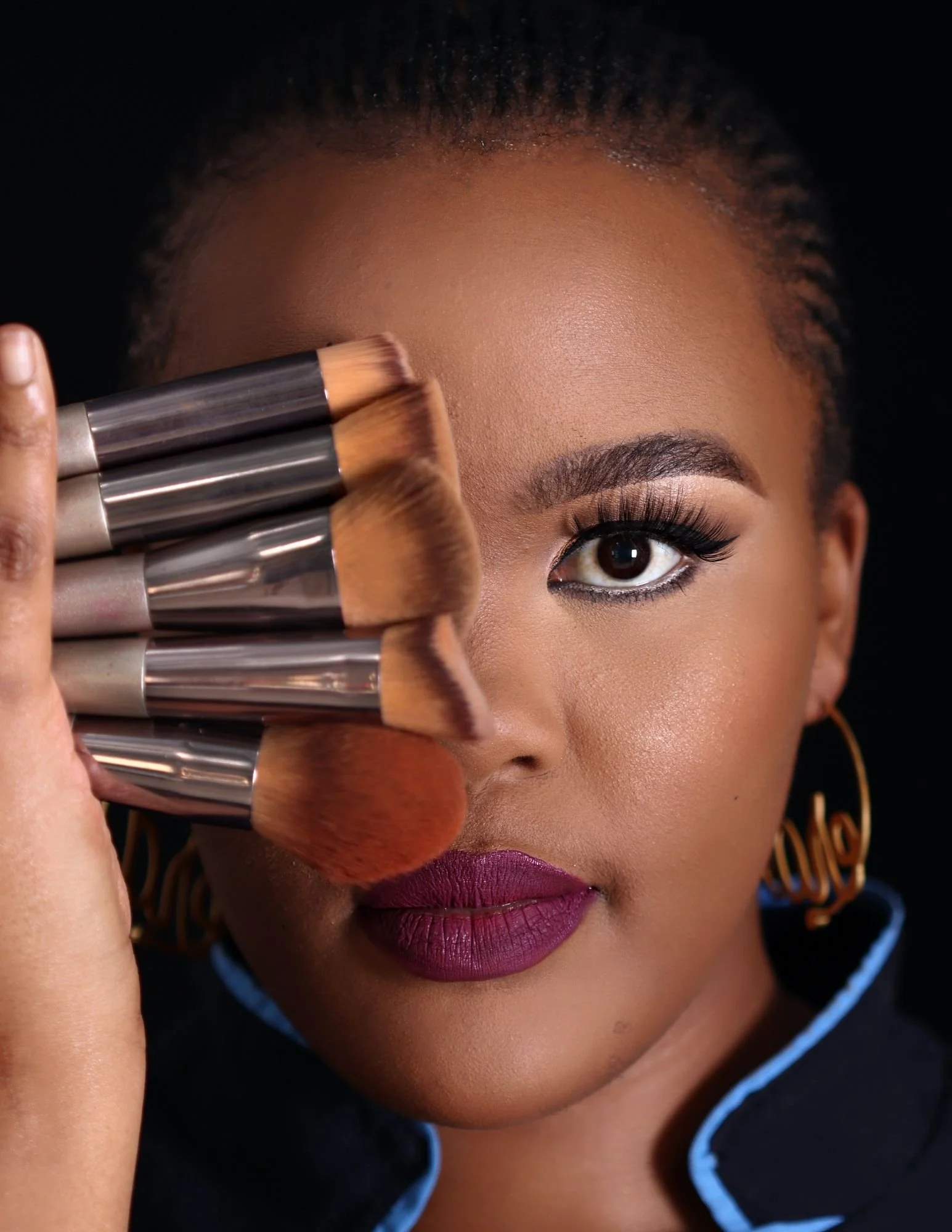 Close-up of a woman holding makeup brushes near her face, with makeup applied including lipstick, eyeliner, and eyeshadow, against a black background.