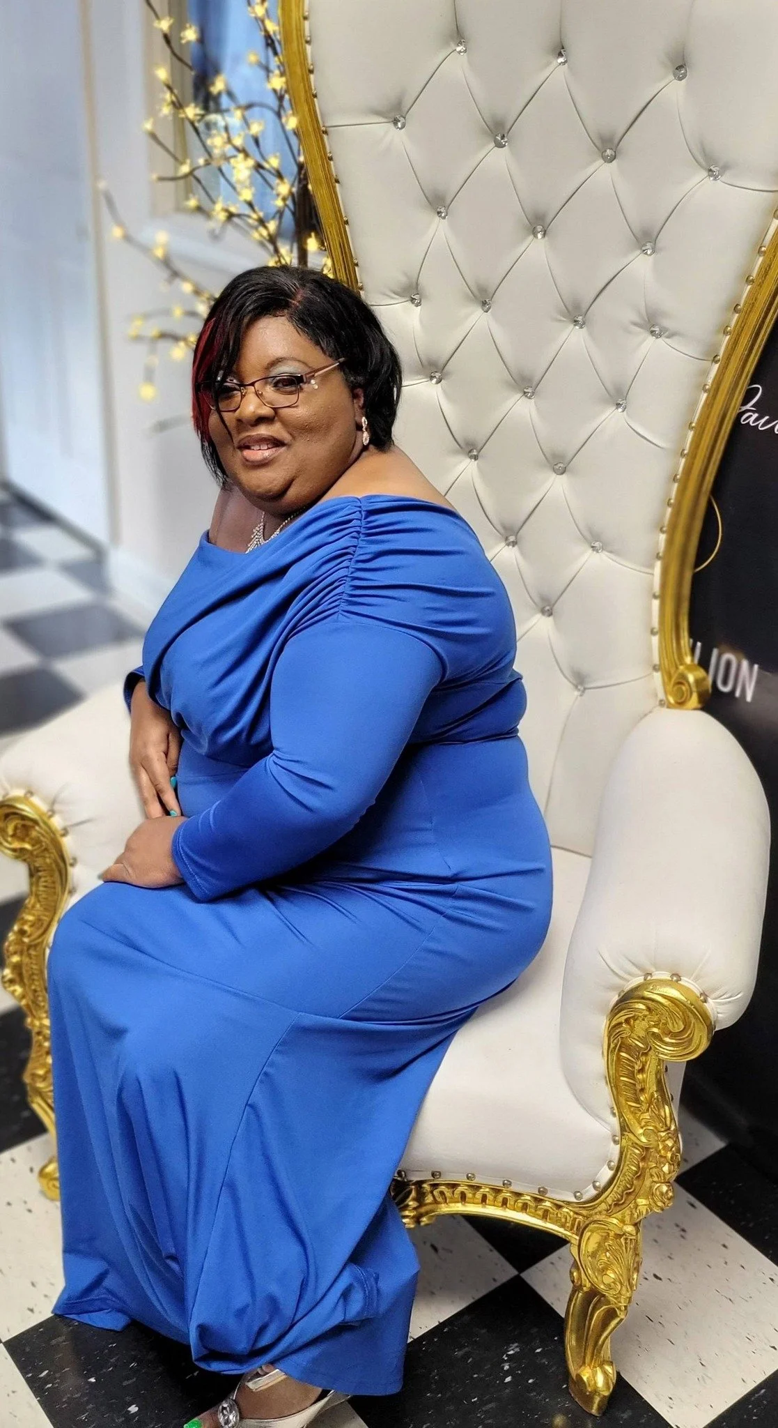 A woman in a blue dress sitting on a white and gold ornate throne chair, indoors with a black and white checkered floor and decorative lighting in the background.