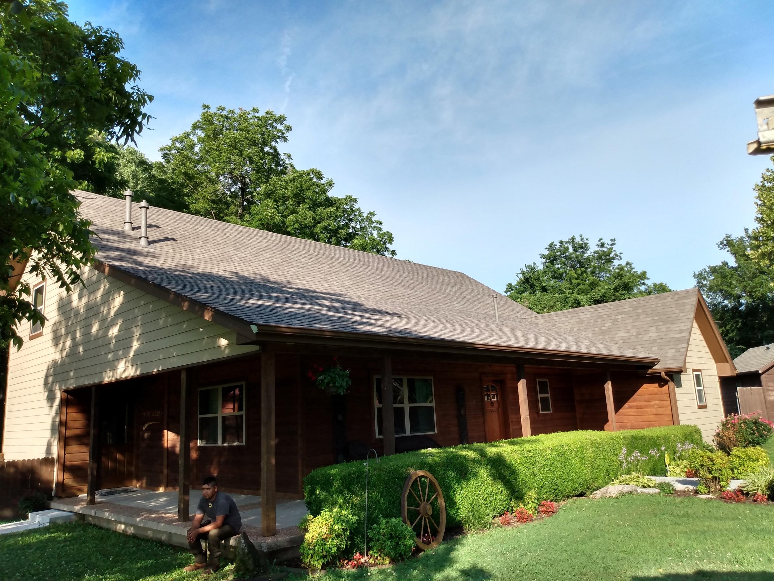 A house with wooden and beige siding, a porch with wooden support beams, and a man sitting on a stone ledge in front, surrounded by trees and a well-kept lawn.