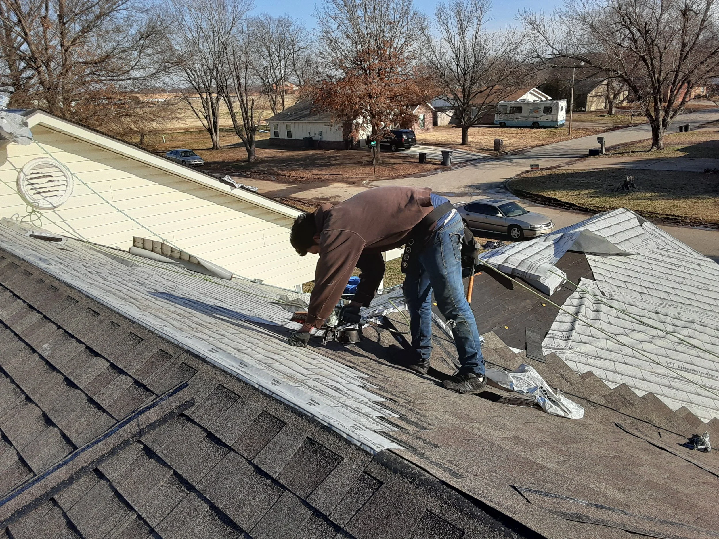 Two workers on a rooftop replacing shingles, with a suburban neighborhood and parked cars in the background during daylight.
