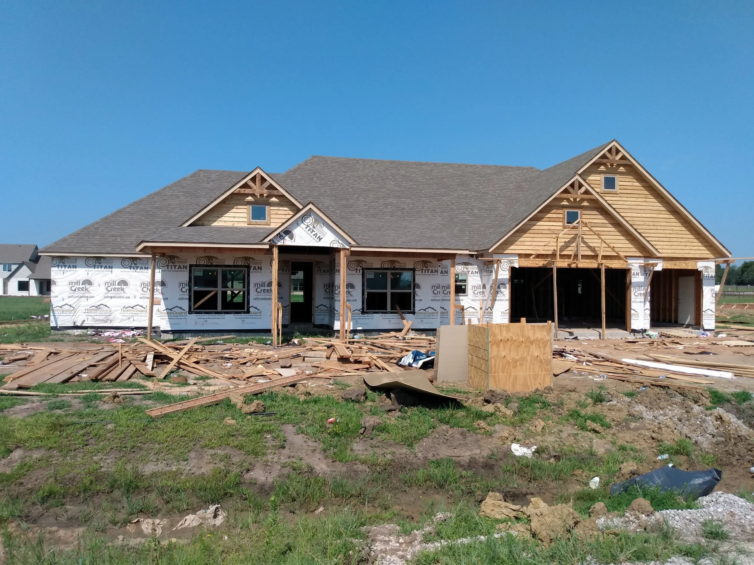 A house under construction with an unfinished facade, surrounded by construction debris, wooden framing, and building materials on a grassy area under a clear blue sky.