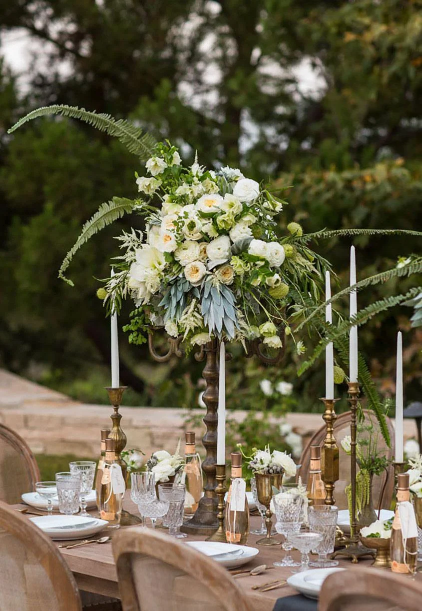 Elegant outdoor dining table decorated with a tall floral centerpiece of white flowers and greenery, surrounded by gold candlesticks, glassware, and plates, on a wooden table with chairs, set in a garden or park area.