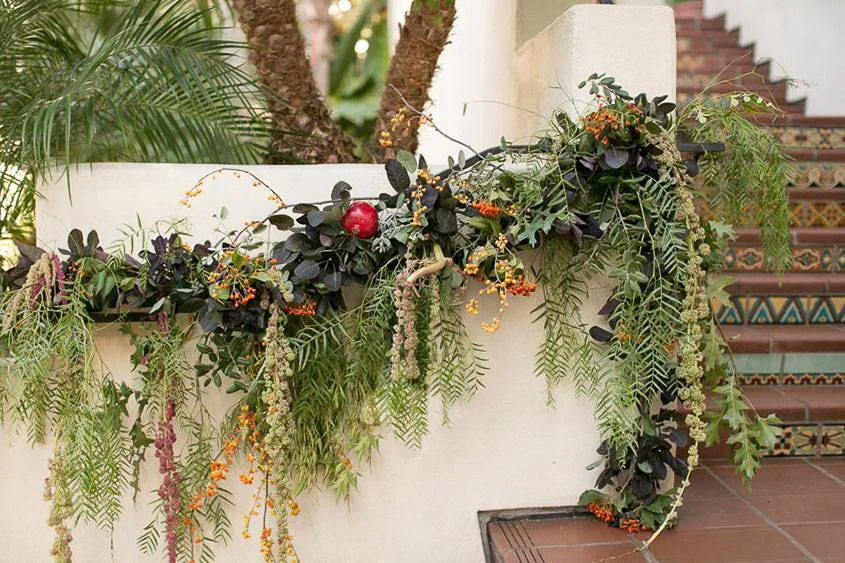 Decorative floral arrangement with greenery, orange berries, and a red apple on a white wall near patterned tiled stairs.