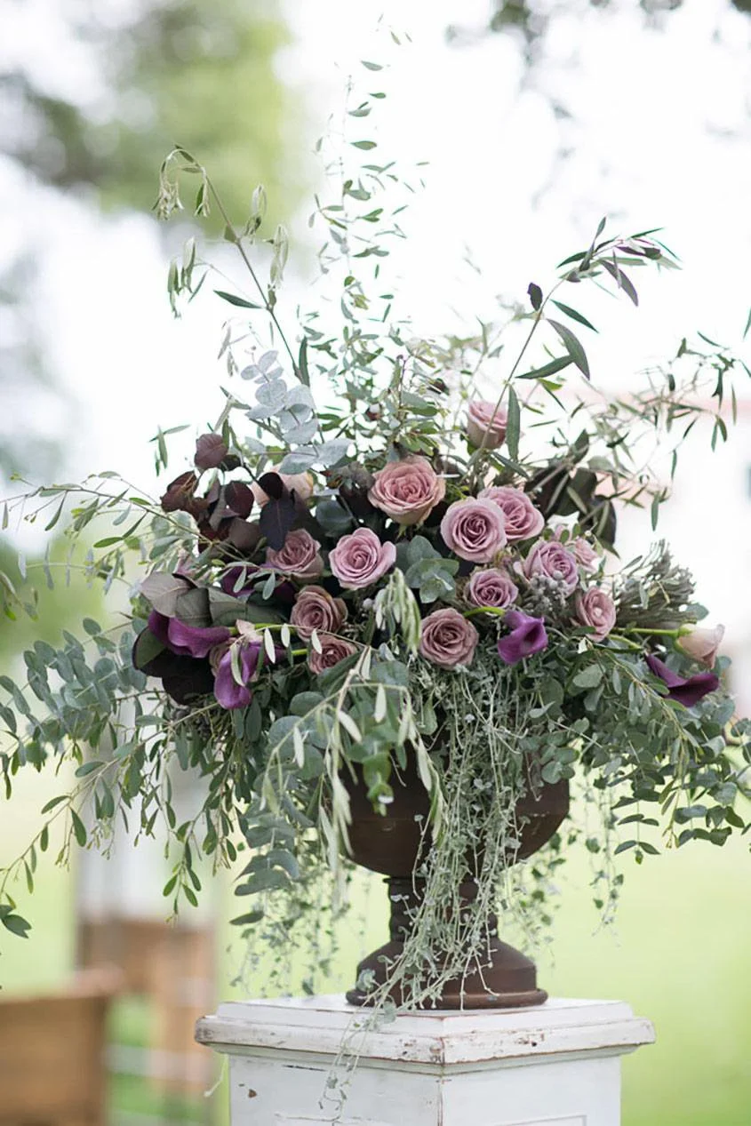 A floral arrangement featuring pink roses, purple calla lilies, and assorted greenery in a vintage-style bronze urn on a distressed white pedestal, set outdoors.
