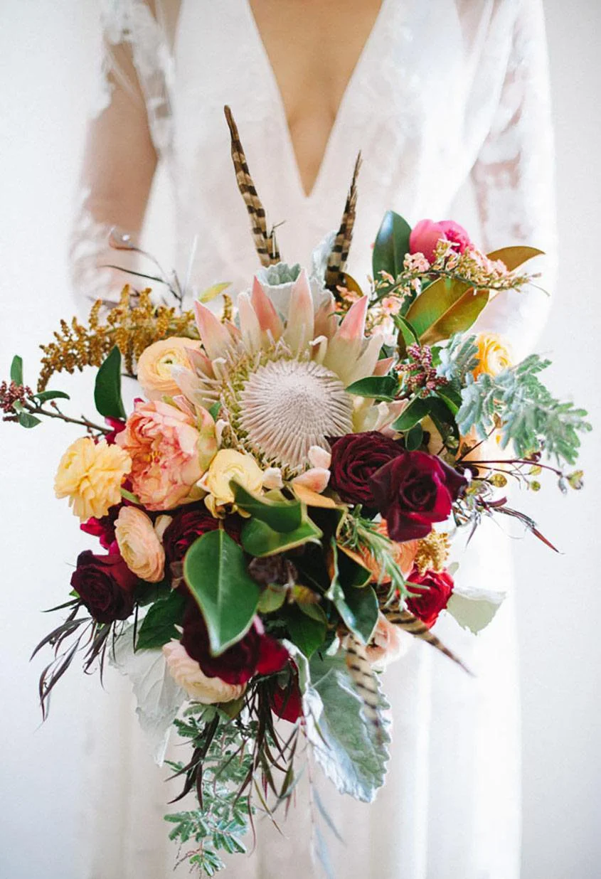 A person in a white lace dress holding a colorful bouquet of flowers, including roses, peonies, and greenery, with feathers and other decorative elements.