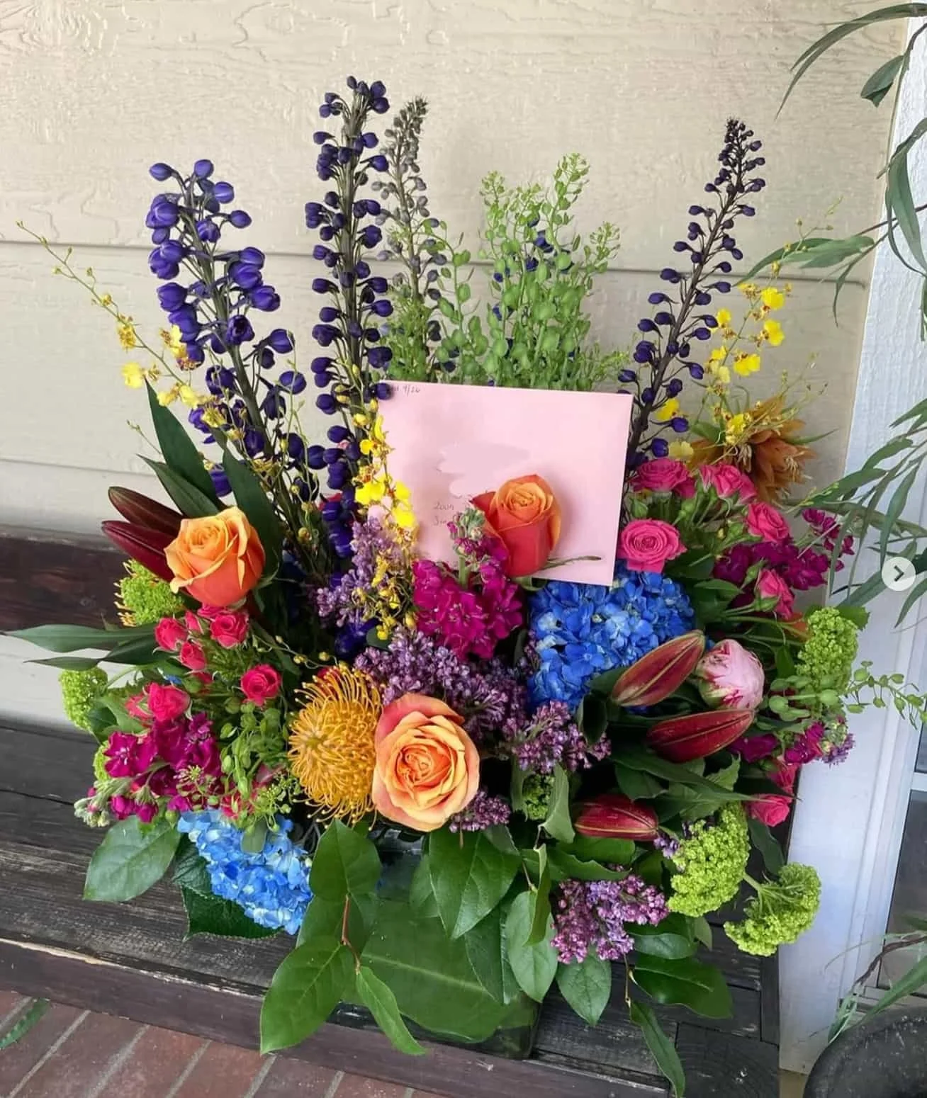 A colorful bouquet of flowers including roses, delphiniums, hydrangeas, and other assorted flowers arranged on a wooden stand against a neutral wall.