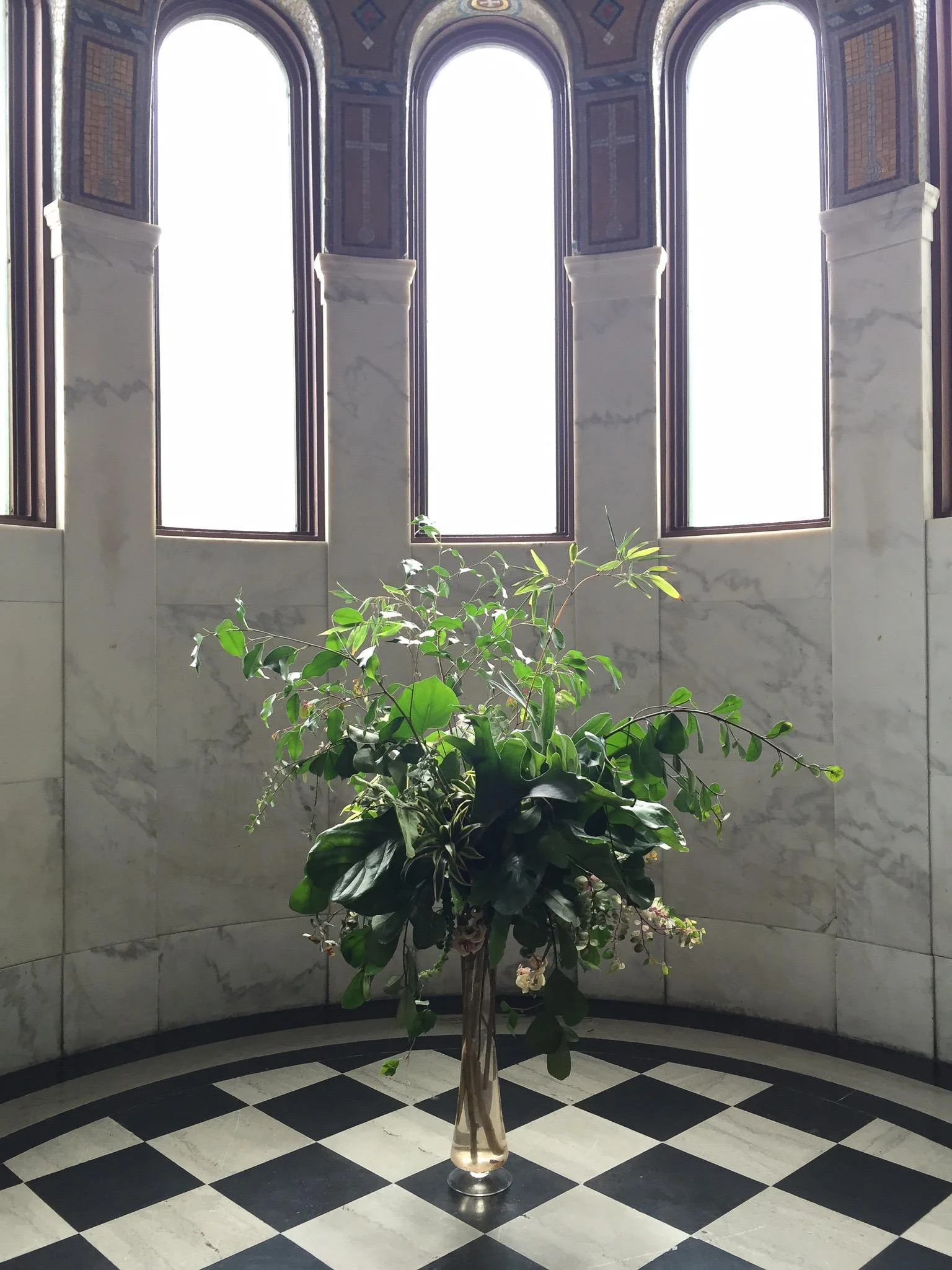 A floral arrangement with green leaves and small white flowers in a tall glass vase, placed on a checkered black and white floor in front of a marble wall with tall, arched windows letting in bright sunlight.
