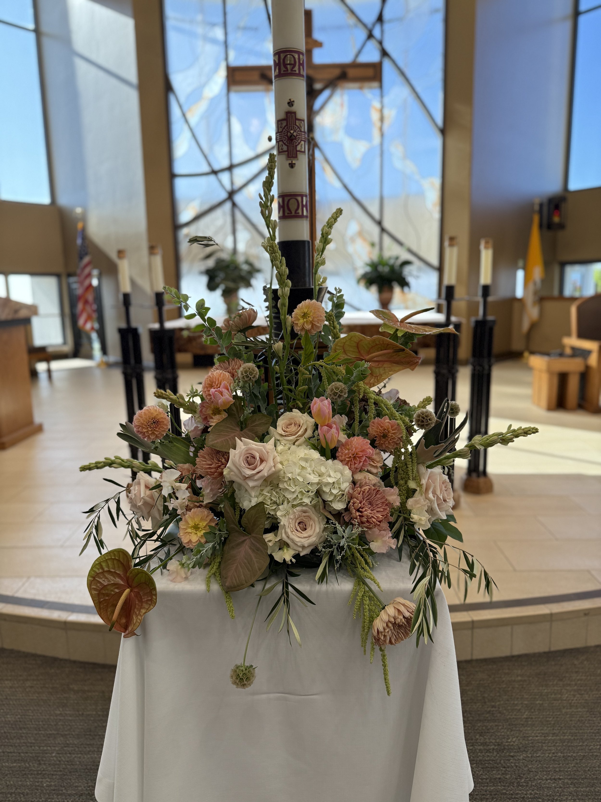 Flowers arranged around a tall Paschal candle with religious symbols inside a church.