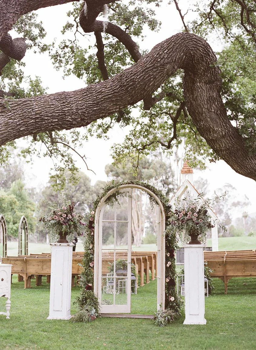 Outdoor wedding arch decorated with flowers and greenery, under a large tree with sprawling branches.