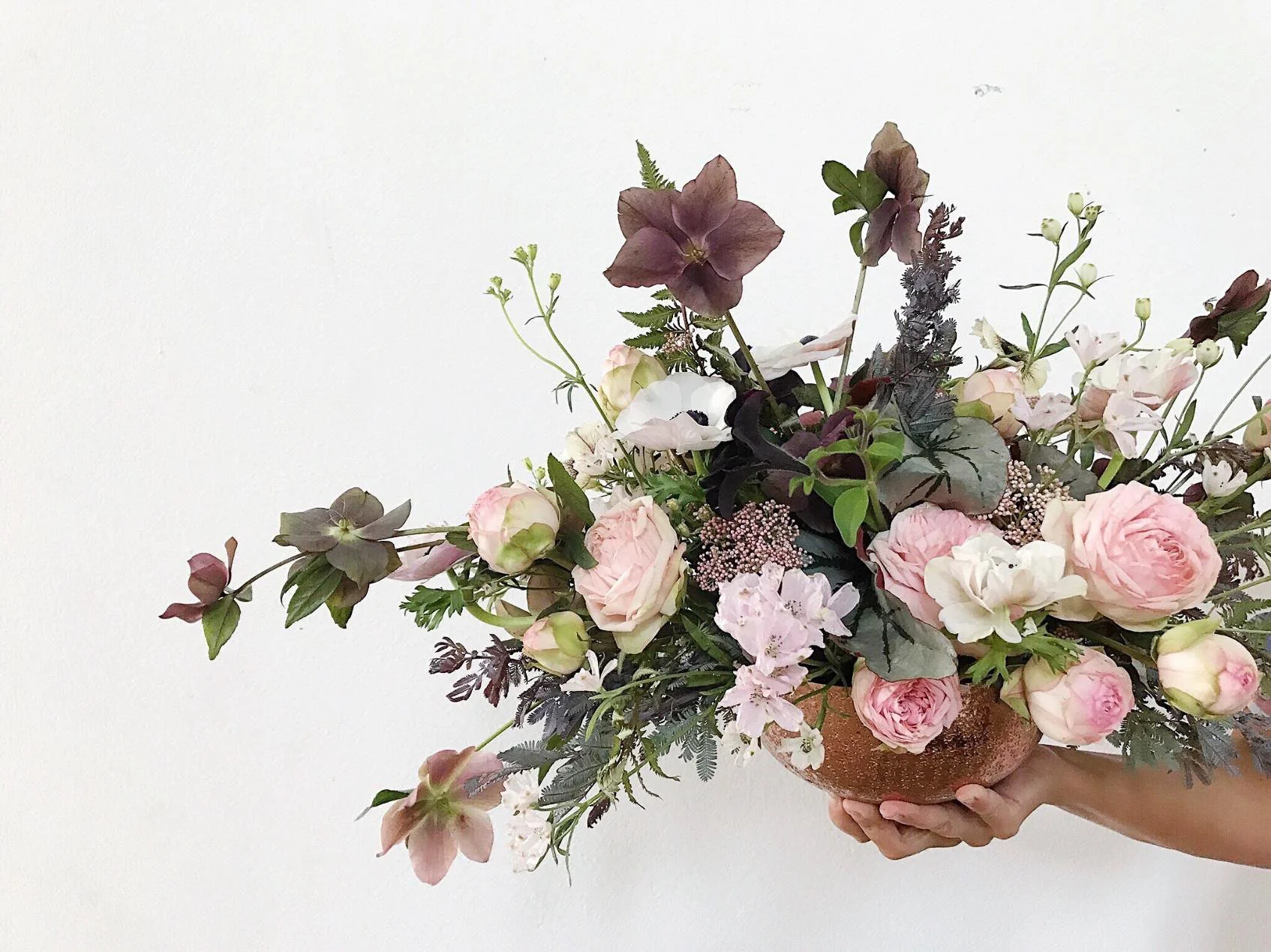 Person holding a large floral arrangement with pink roses, lavender, and greenery in a brown pot against a plain white wall.