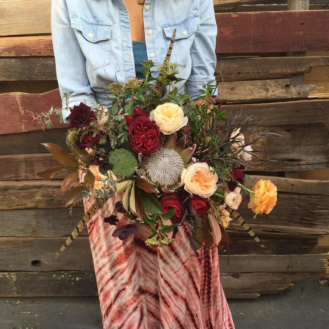 A person holds a large, colorful bouquet of flowers in front of a wooden fence. The person wears a light blue denim shirt and a red and white patterned skirt.