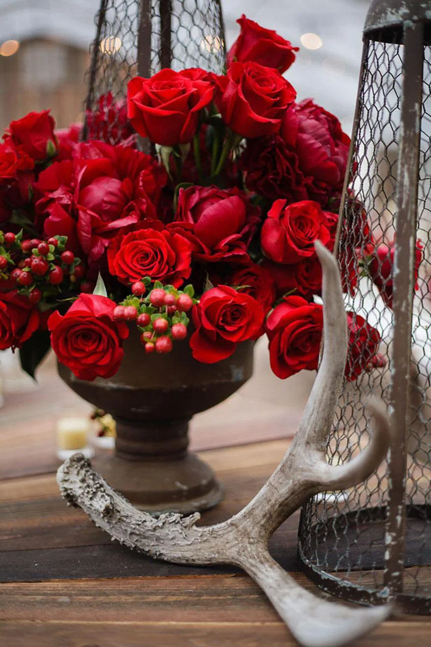 A bouquet of red roses and berries in a black vase, with an antler and a wire decorative cage on a wooden surface.