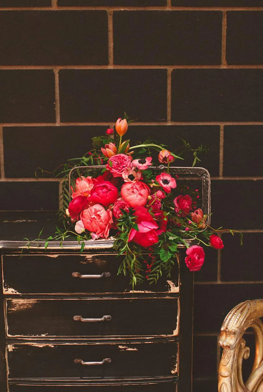 A vintage black chest of drawers with peeling paint and three drawers, topped with a colorful arrangement of pink and red flowers, against a dark brick wall.