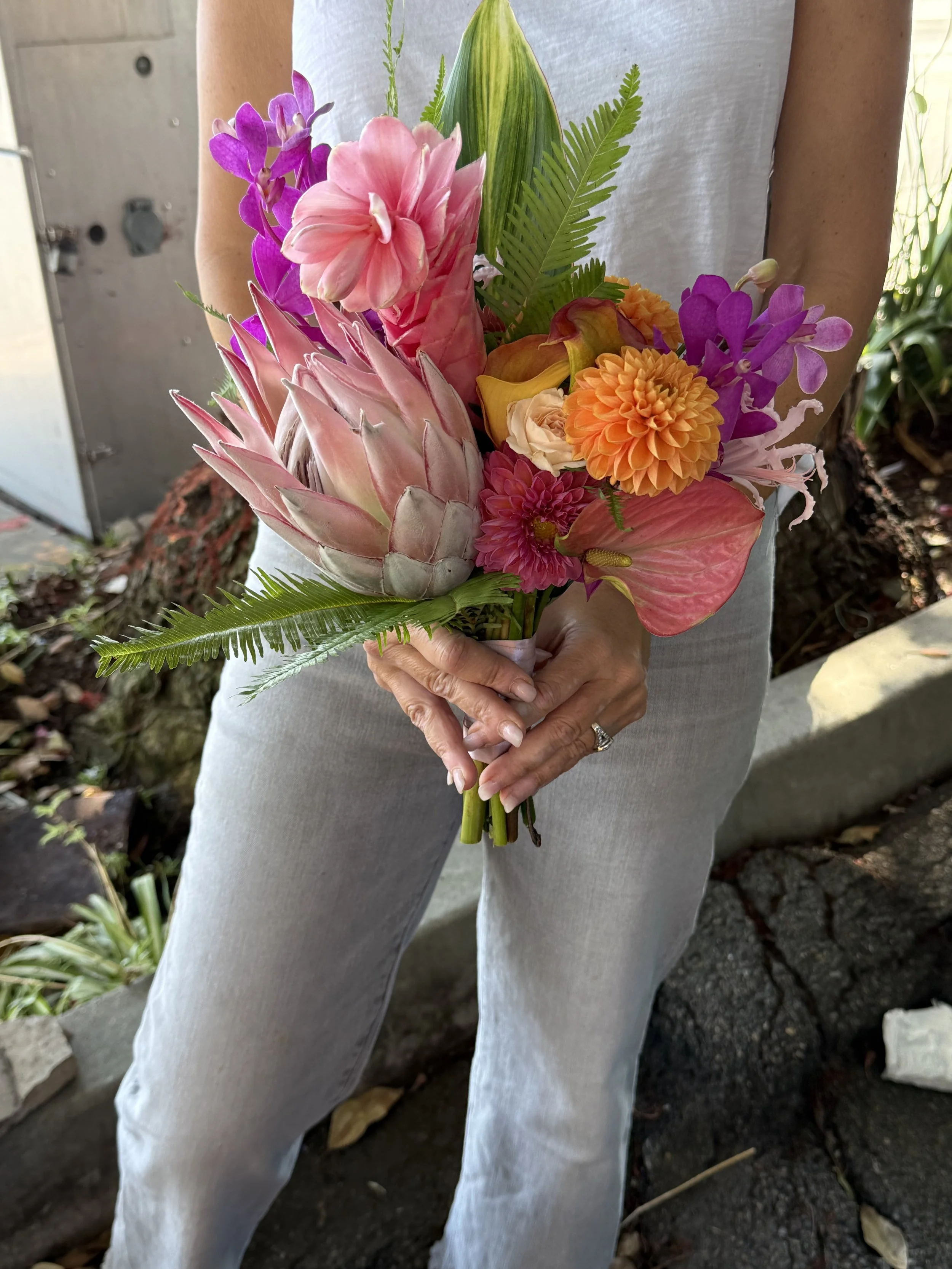 A person holding a colorful bouquet of tropical flowers, including pink protea, pink dahlias, orange dahlias, purple orchids, and green foliage. The person is wearing a white shirt and light-colored pants, standing outdoors.