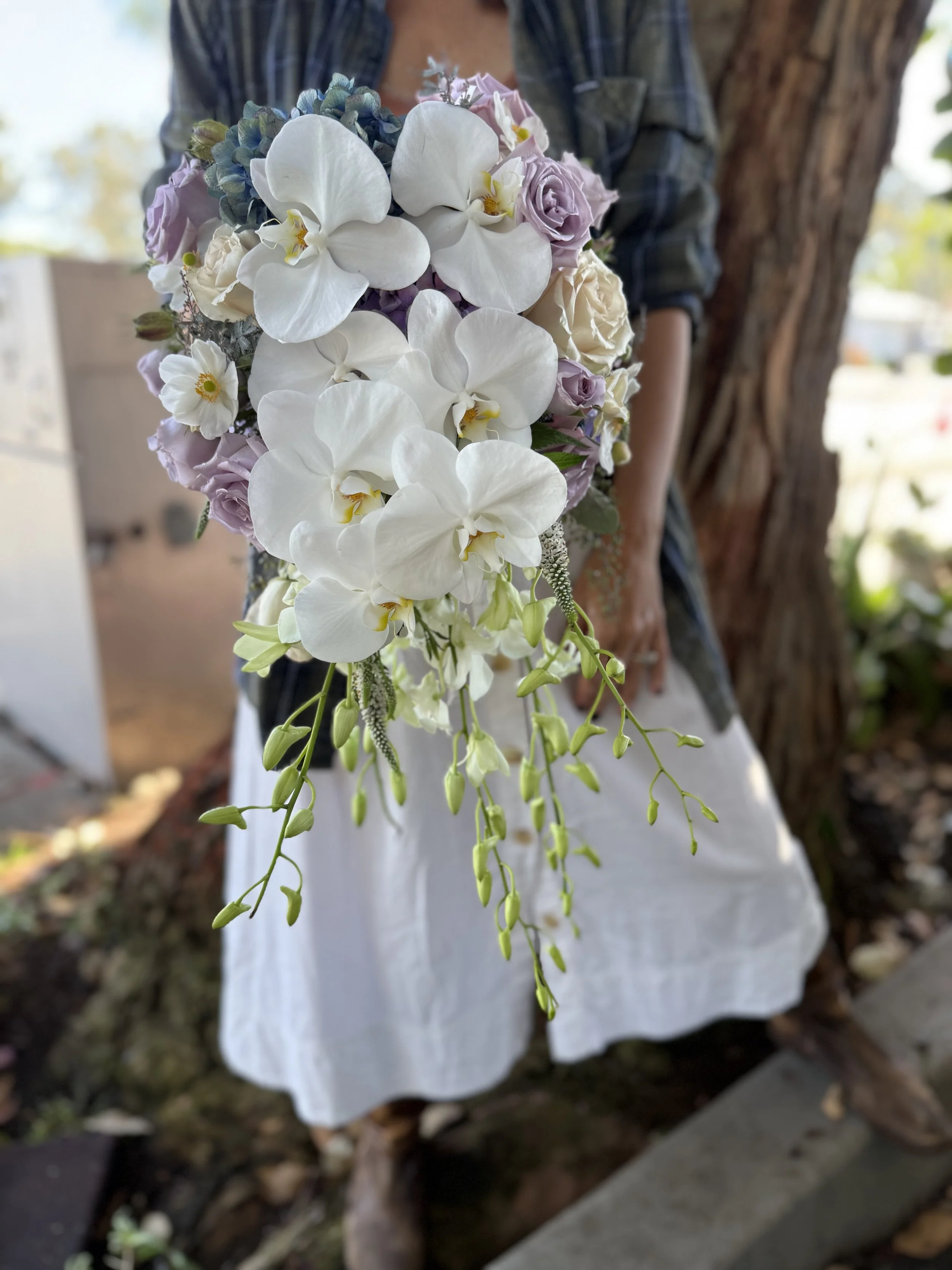 A person holding a cascading bouquet of white orchids, lavender roses, and other pastel flowers, standing outdoors near a tree.