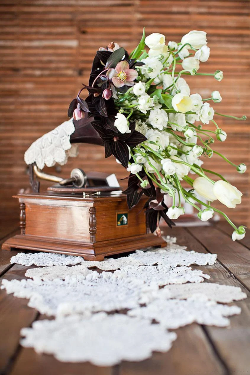 A vintage gramophone with a large bouquet of white and pink flowers, including white roses and dark purple leaves, placed on a crochet doily on a wooden table, with a wooden wall in the background.