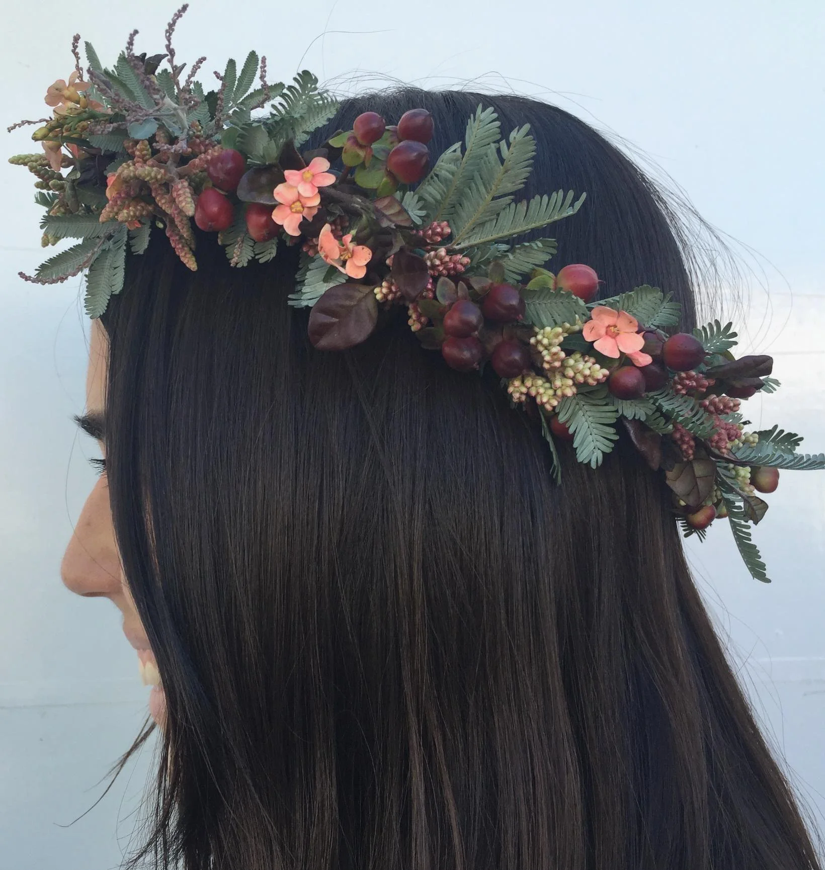 A woman with long dark hair wearing a floral headband with greenery, berries, and small pink flowers.