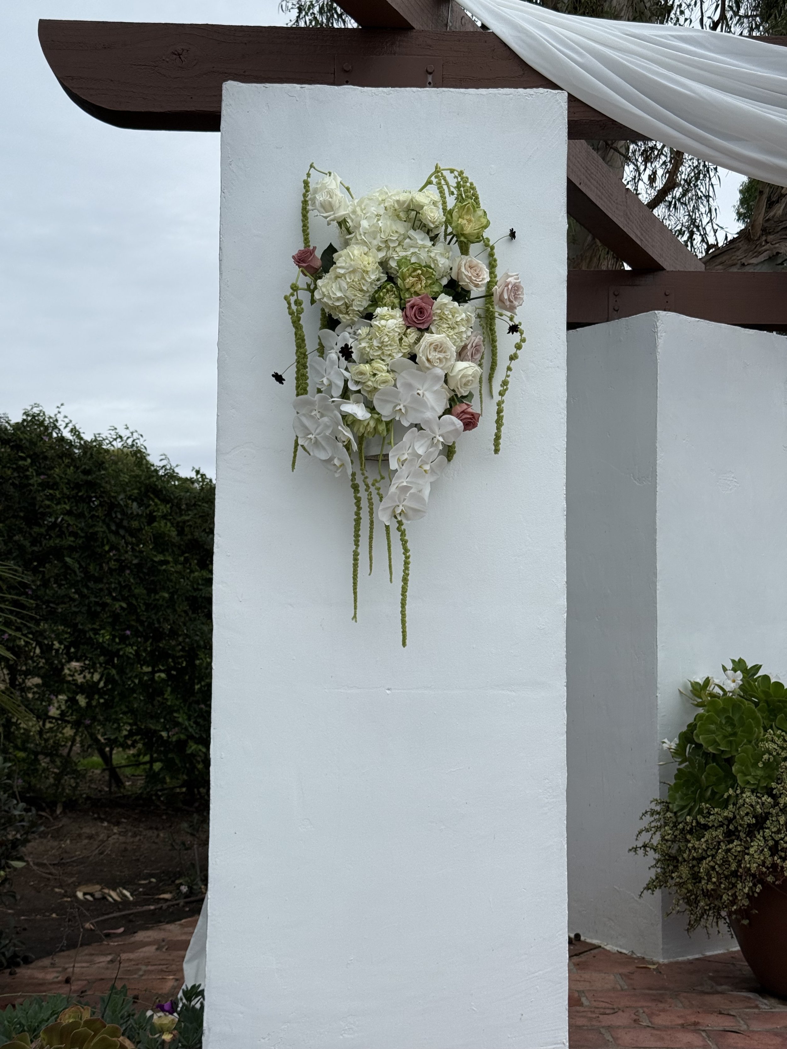 A floral arrangement featuring white hydrangeas, white orchids, pink roses, and hanging green amaranthus, attached to a white wall or pillar at an outdoor setting.