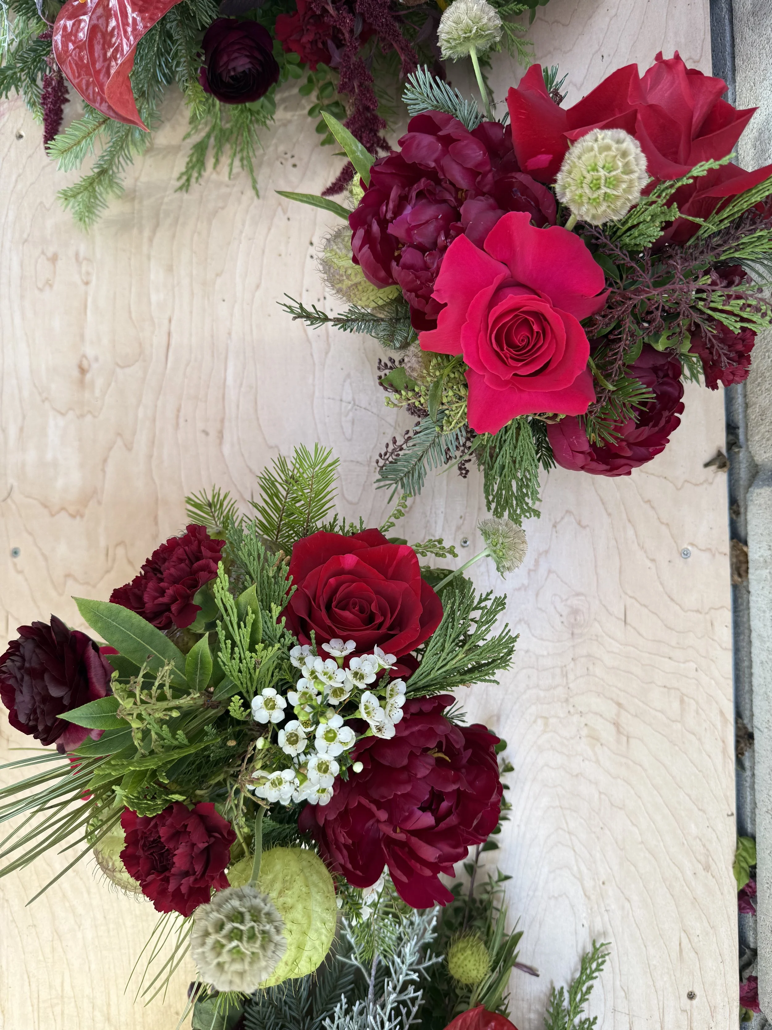 Red and burgundy floral arrangement with roses, carnations, and greenery on a light wooden background.