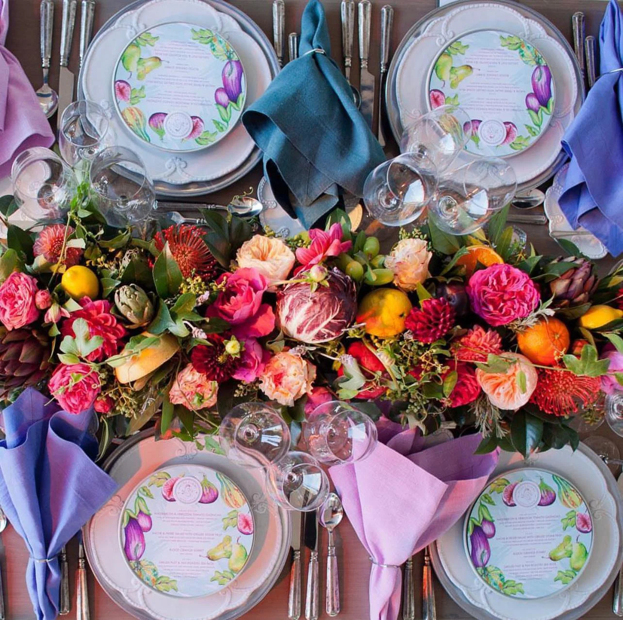 A colorful table setting with plates, glasses, and napkins, and a vibrant floral centerpiece featuring pink, red, orange, and peach flowers with green foliage.