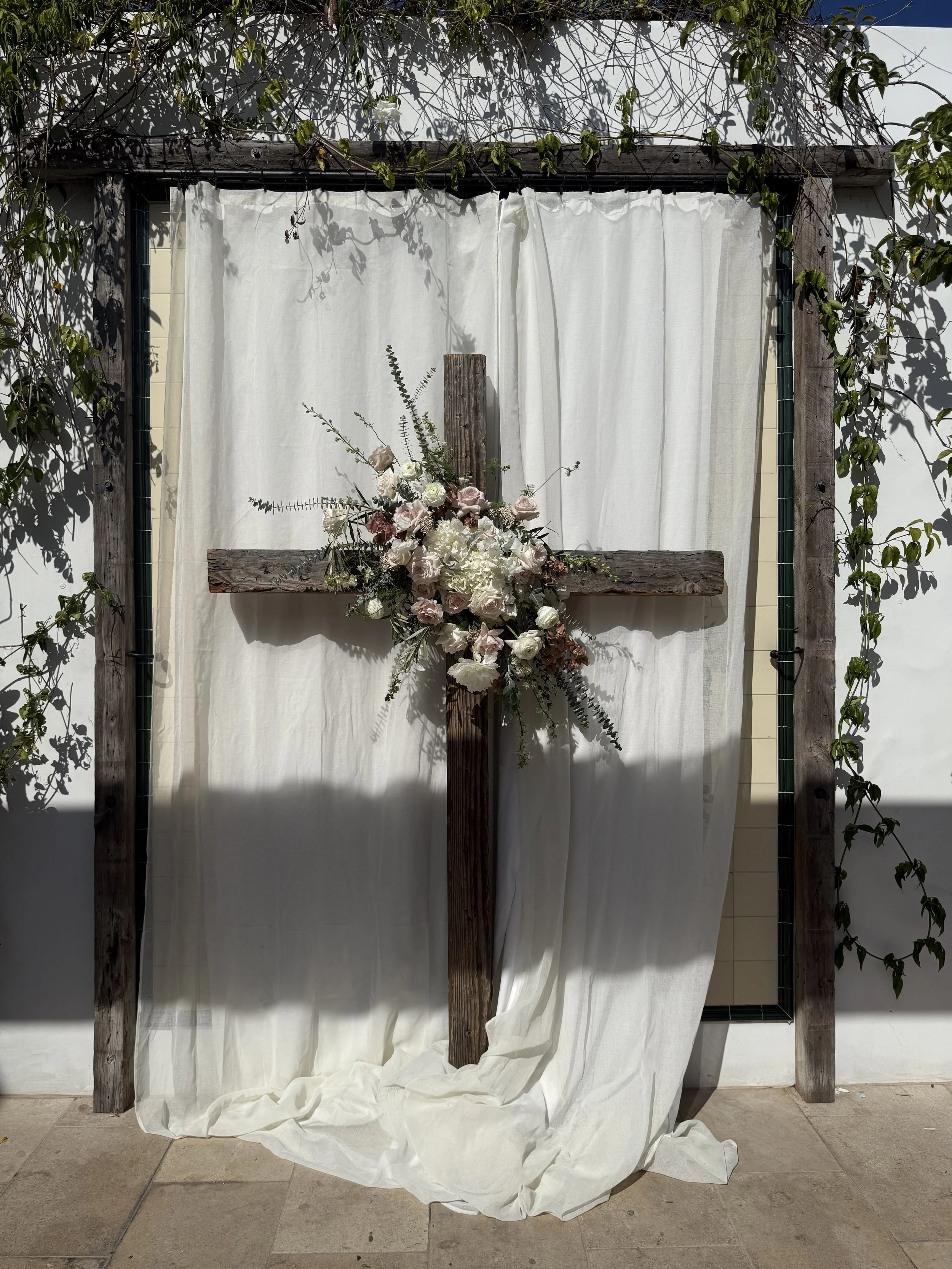Wooden cross decorated with white and blush pink roses and greenery, positioned in front of a white fabric backdrop with a wooden frame, outdoors with shadows and greenery.