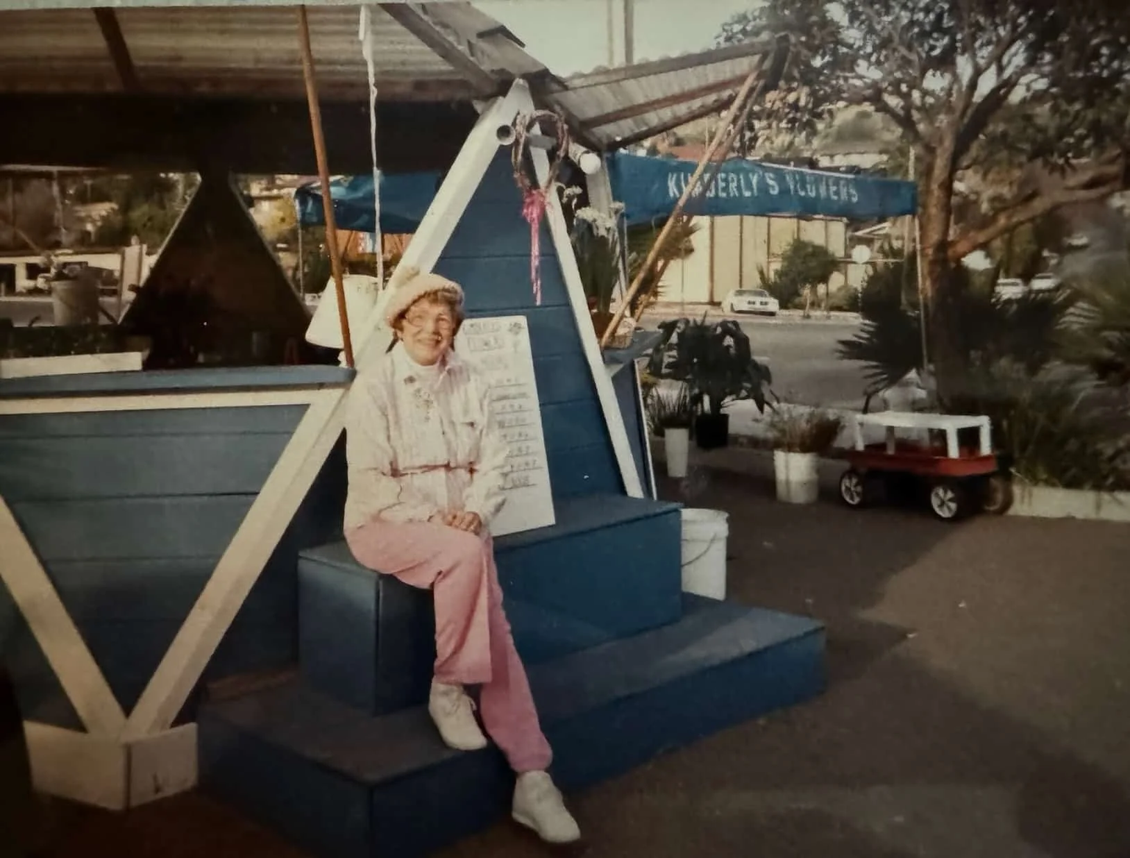 A woman with glasses and a hat sitting on blue steps outside a store named 'Kenderly's Flowers.' She is wearing a pink pair of pants, a white shirt, and white shoes, smiling at the camera. Behind her, there is a sign and some potted plants, with a street and parked cars in the background.