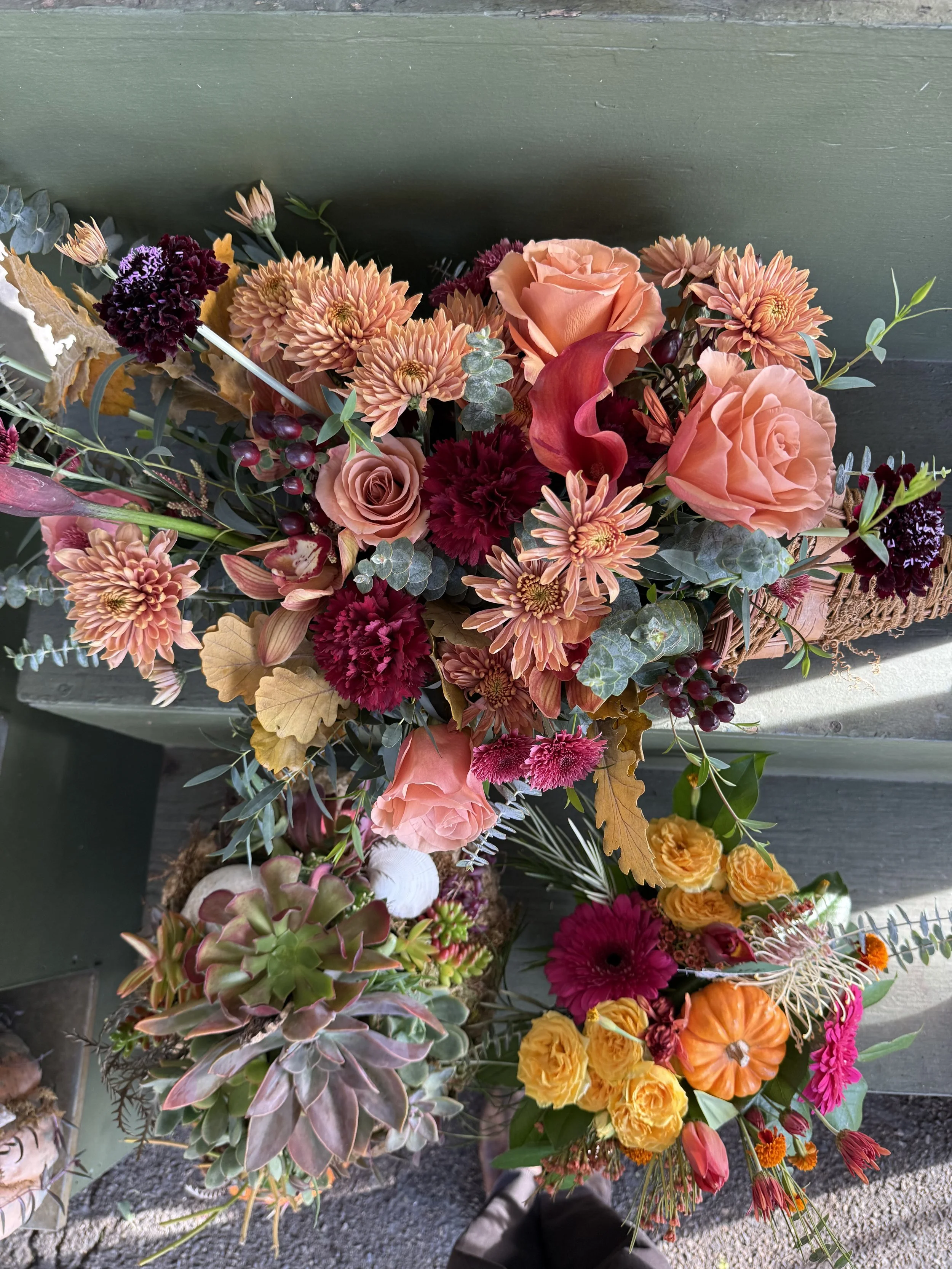 A colorful bouquet of flowers on a dark wooden surface, including roses, chrysanthemums, carnations, dahlias, coxcombs, eucalyptus, succulents, pumpkins, and autumn leaves.