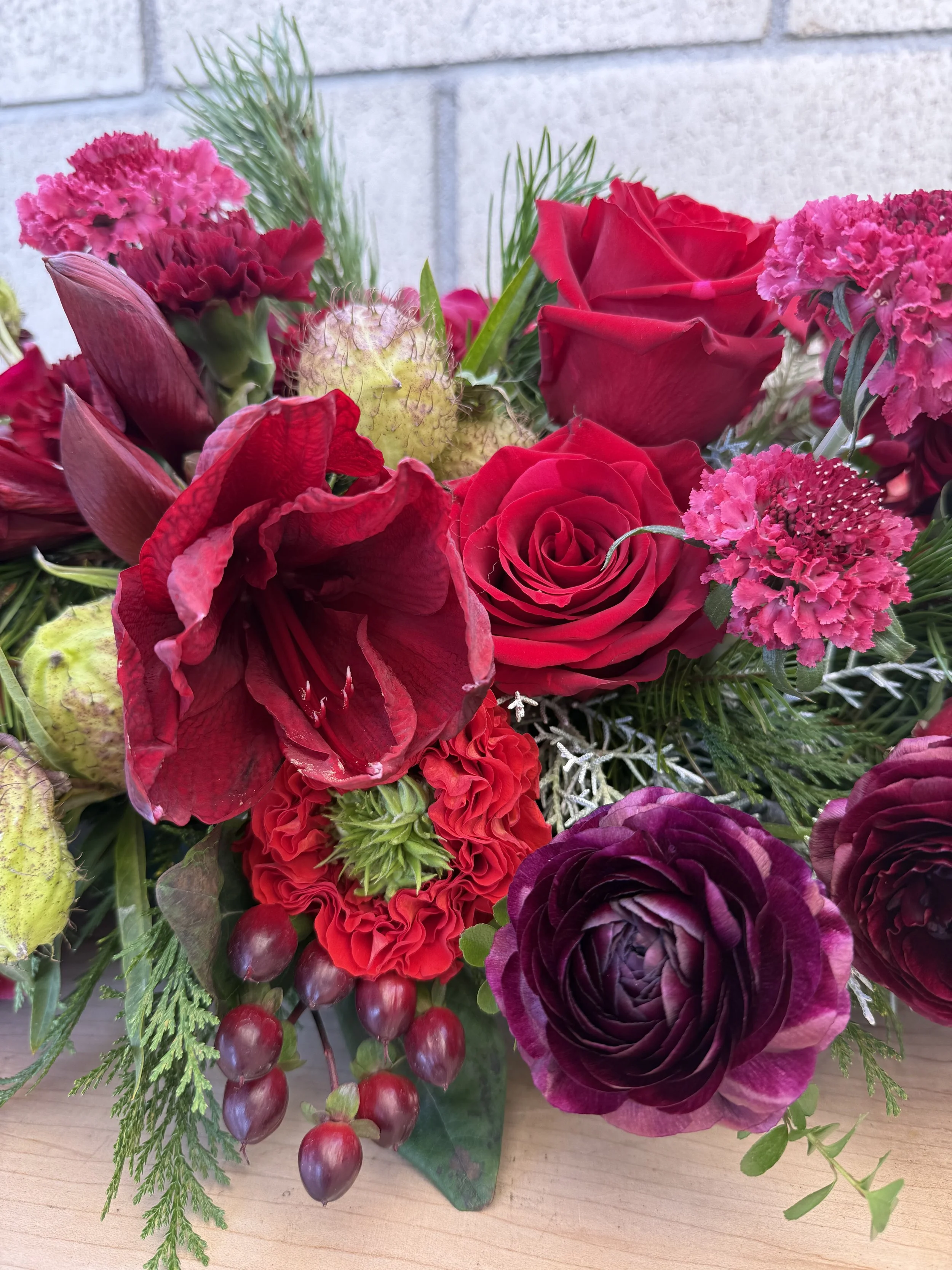 A vibrant bouquet of red roses, pink carnations, purple ranunculus, and red berries on a wooden surface with a brick wall background.