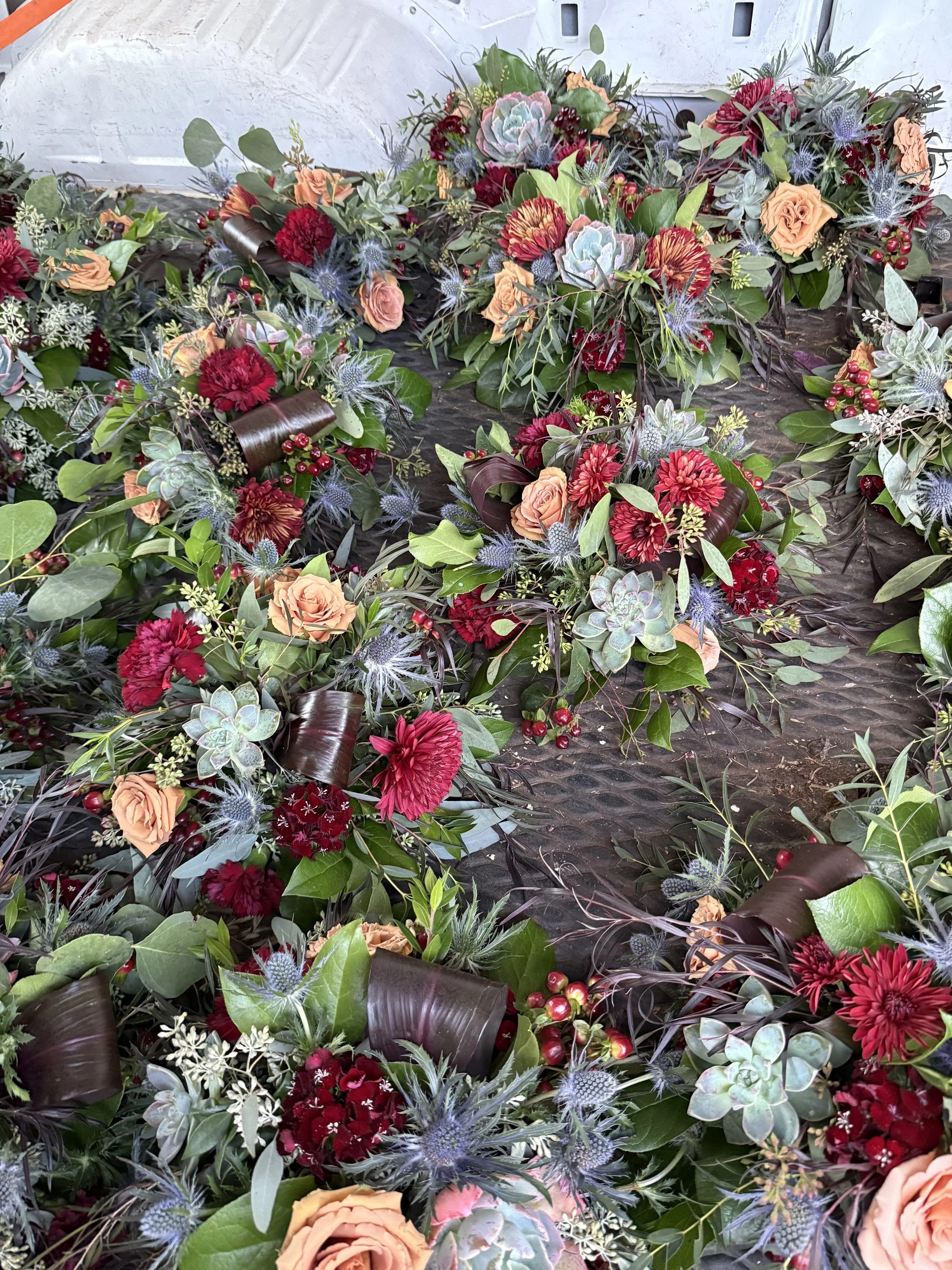 A floral arrangement with pink roses, red carnations, orange chrysanthemums, succulents, and green foliage, arranged in a circular pattern on a dark surface.