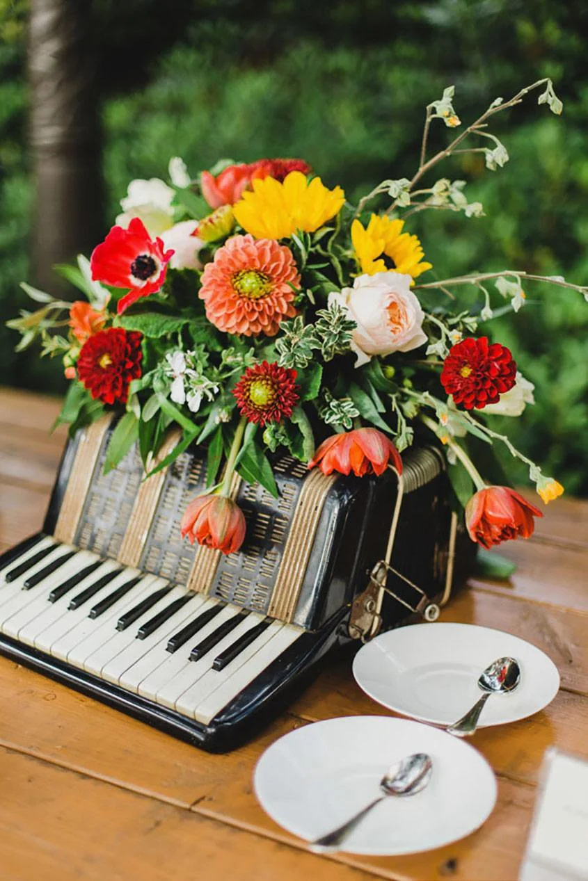 A bouquet of colorful flowers arranged on a small black toy piano, with two white plates and spoons on a wooden table.