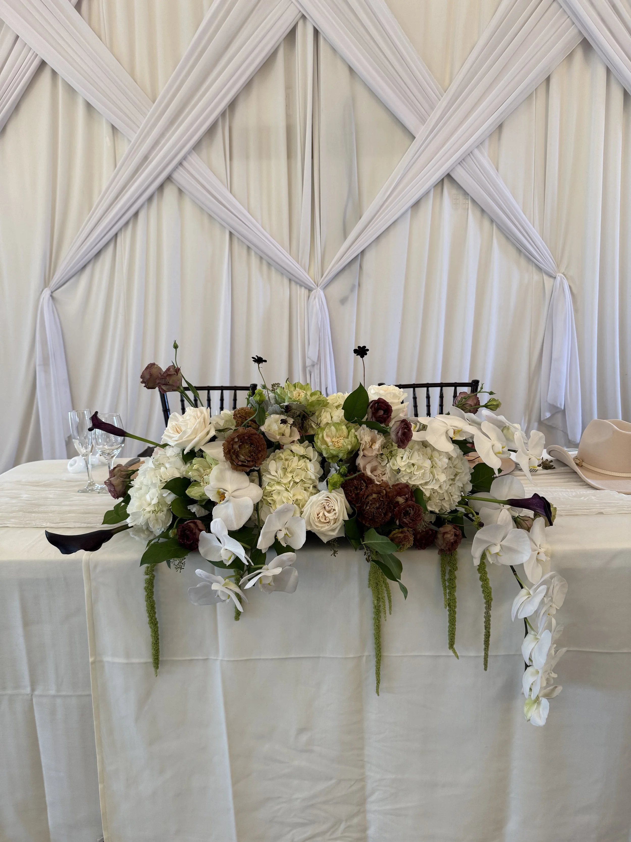 Elegant wedding table setup with a large floral centerpiece of white and dark red flowers, surrounded by champagne glasses, with an elaborate white draped backdrop.