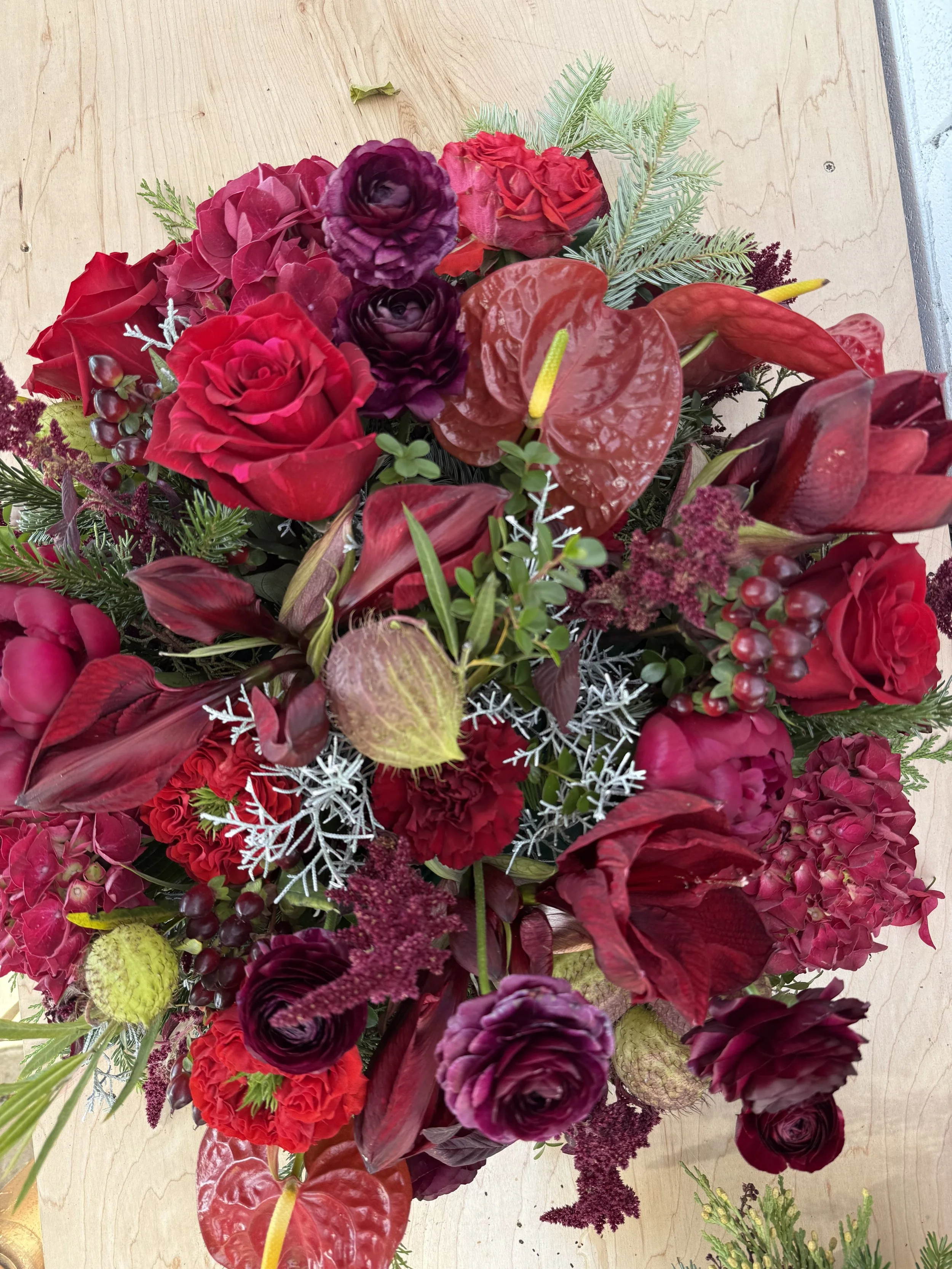 A vibrant floral arrangement with red roses, purple ranunculus, red anthuriums, green succulents, and various greenery, displayed on a light wooden surface.