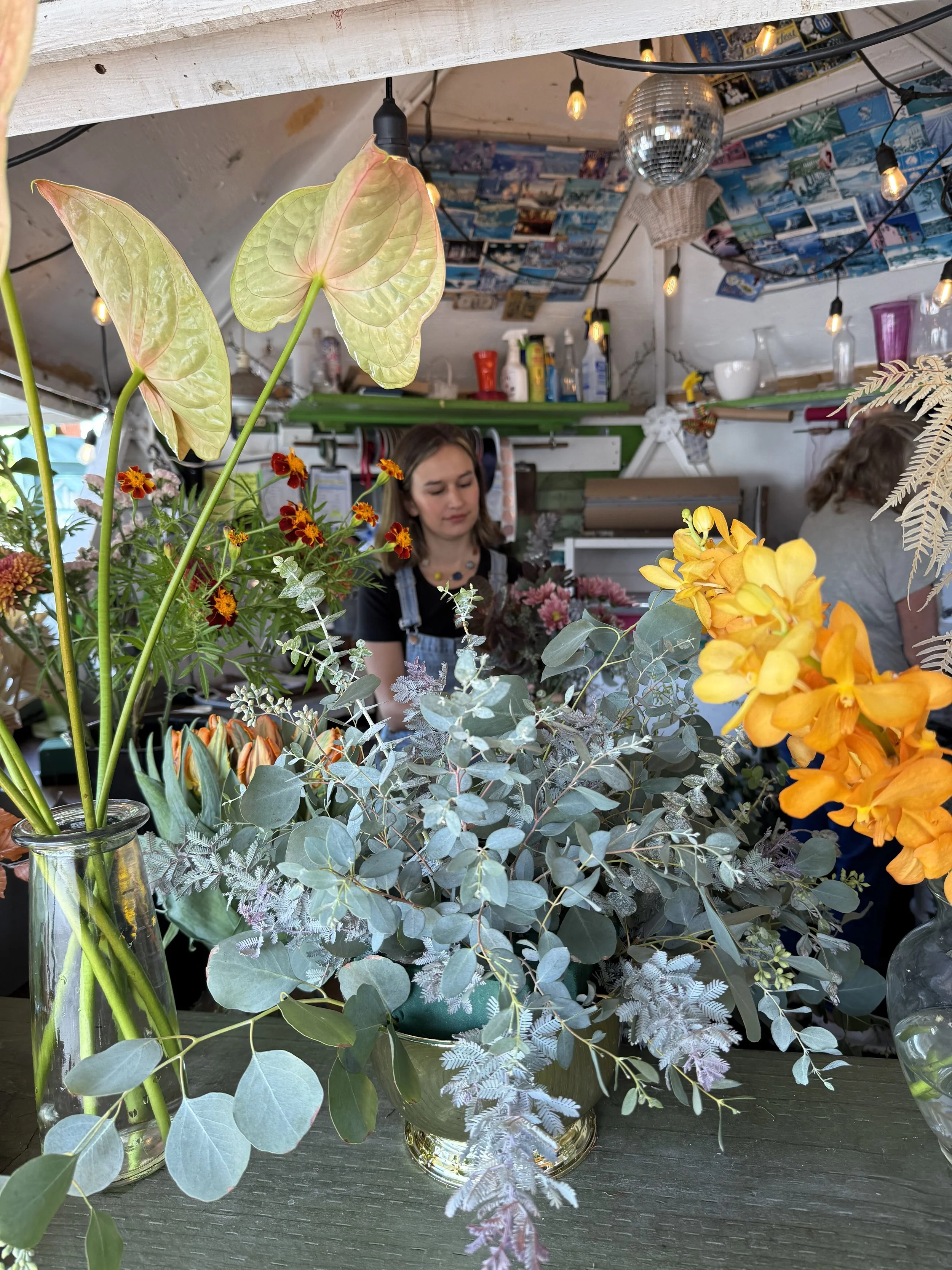 Arrangement of various flowers and greenery including yellow orchids, eucalyptus, and anthuriums in glass vases on a table at a flower shop. In the background, a woman is working among other floral displays with a decorated ceiling and string lights.