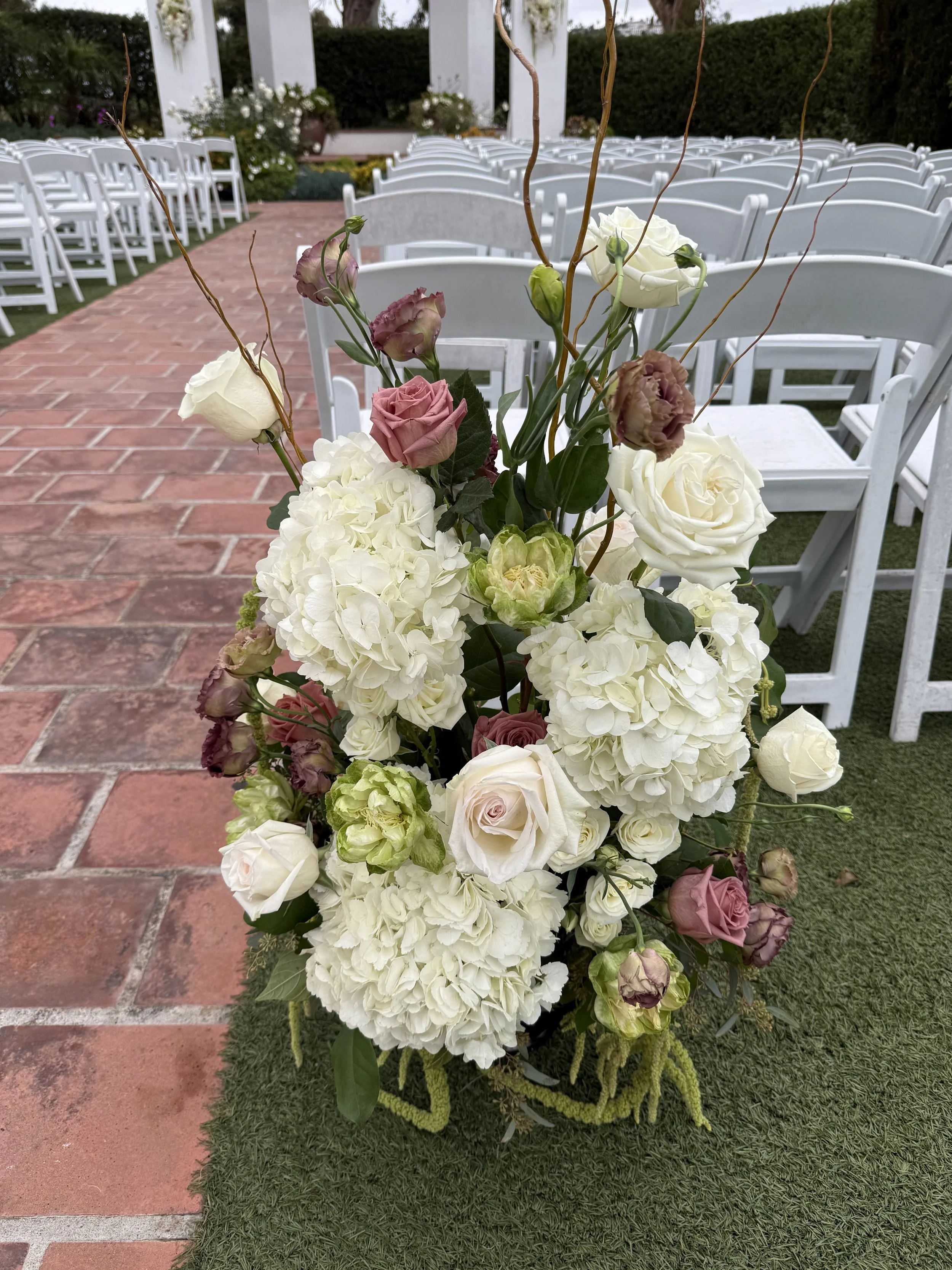A floral arrangement with white roses, hydrangeas, and pink roses on a table in an outdoor wedding setting.