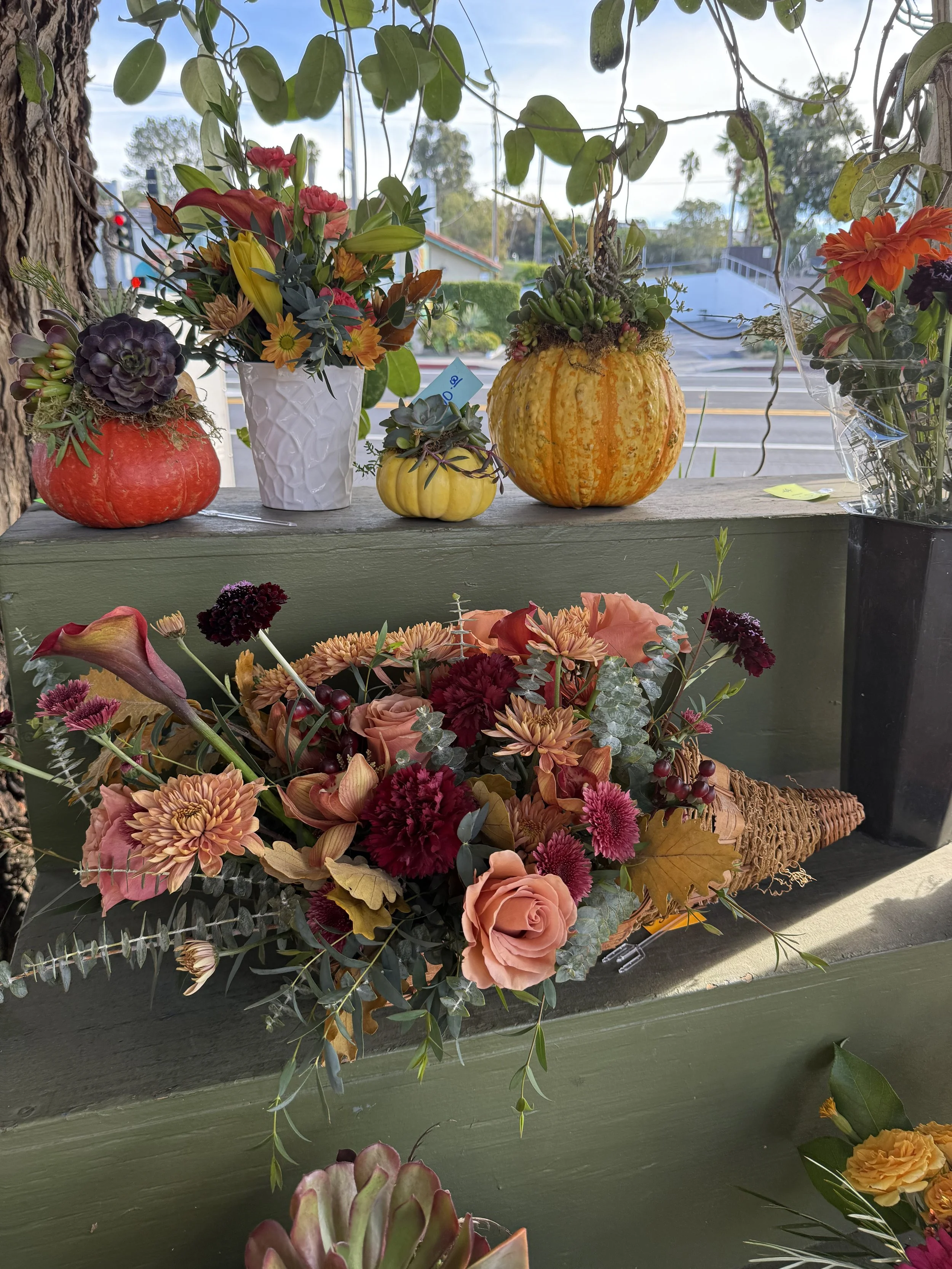 A display of various flowers arranged in bouquets and vases on a green outdoor shelf, with decorative gourds and pumpkins among the flowers.
