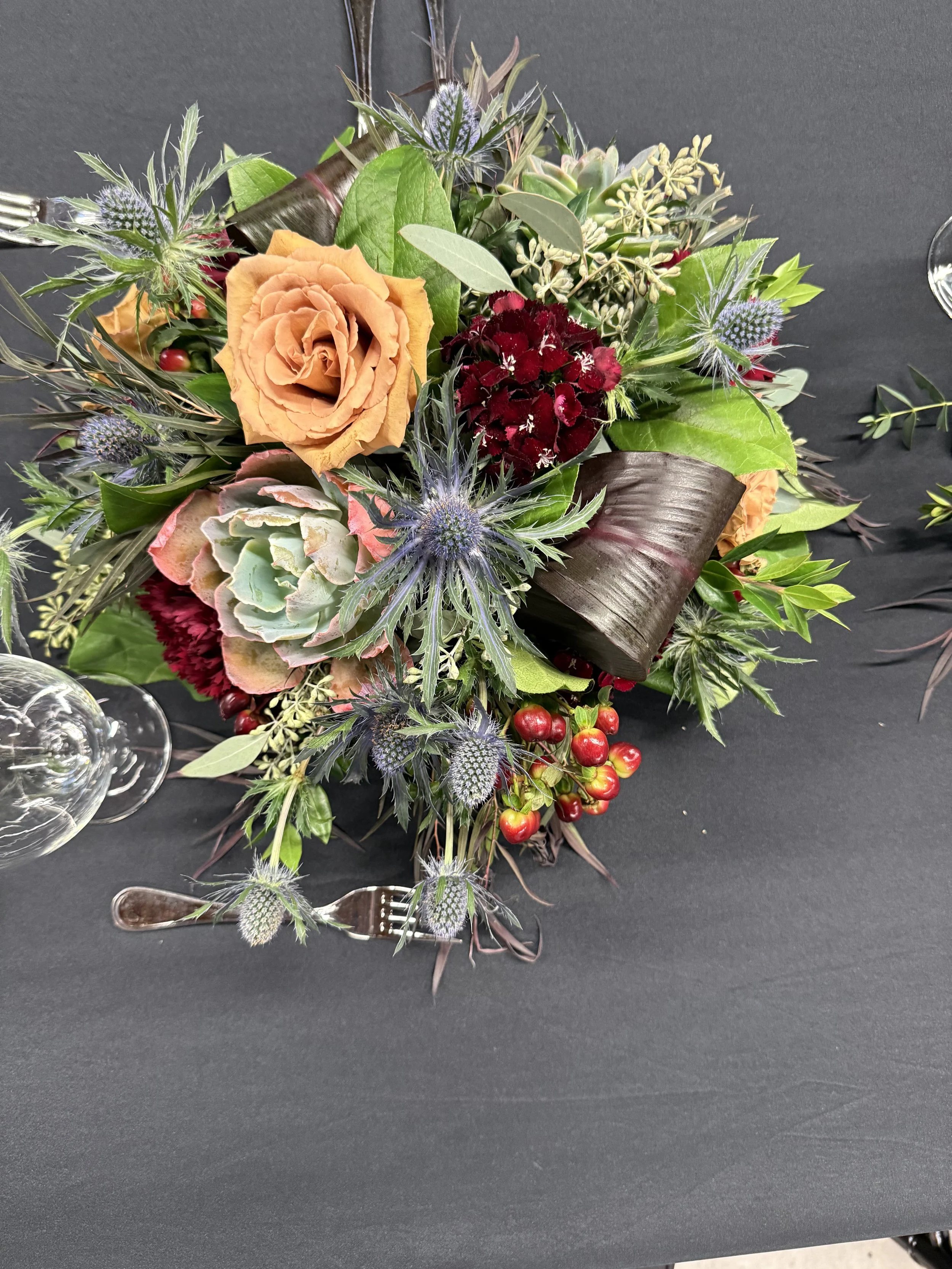 Elegant floral centerpiece on black tablecloth with orange roses, purple thistles, succulents, red berries, and assorted greenery.