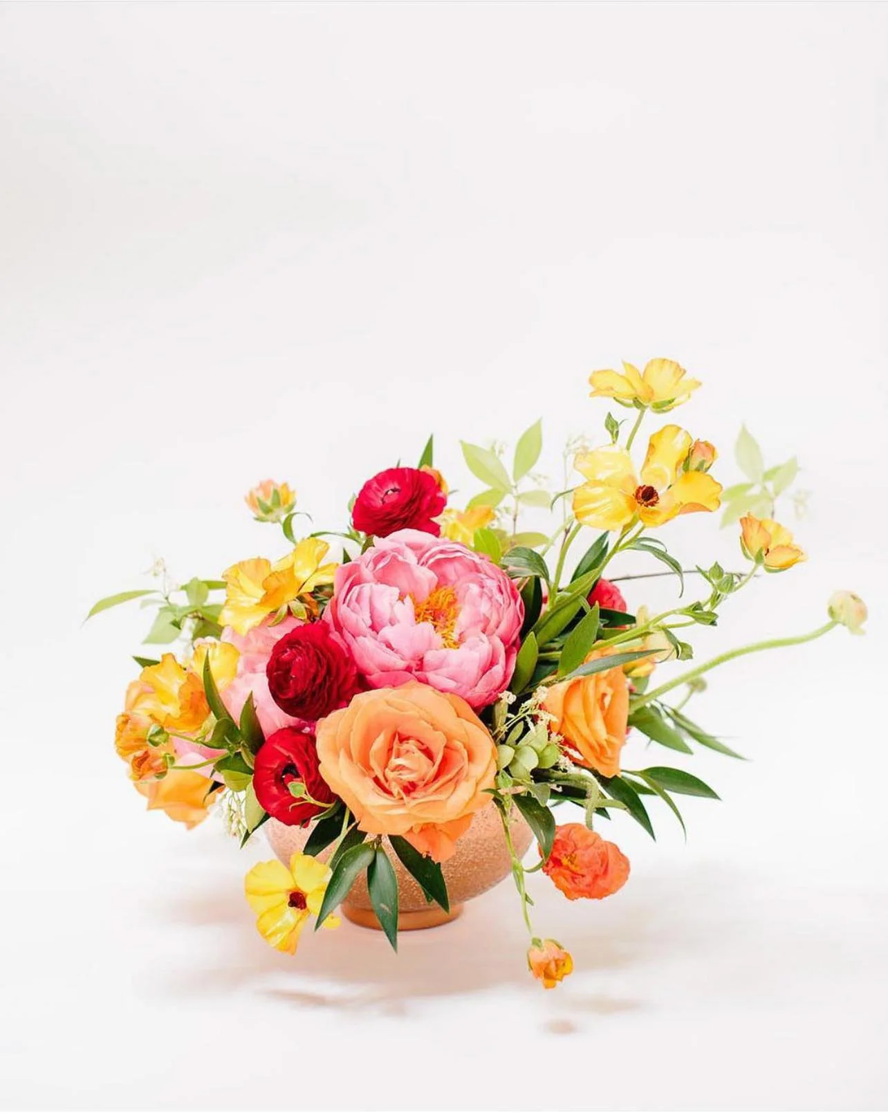 Colorful flower arrangement with pink, orange, yellow, and red flowers in a round vase against a white background.