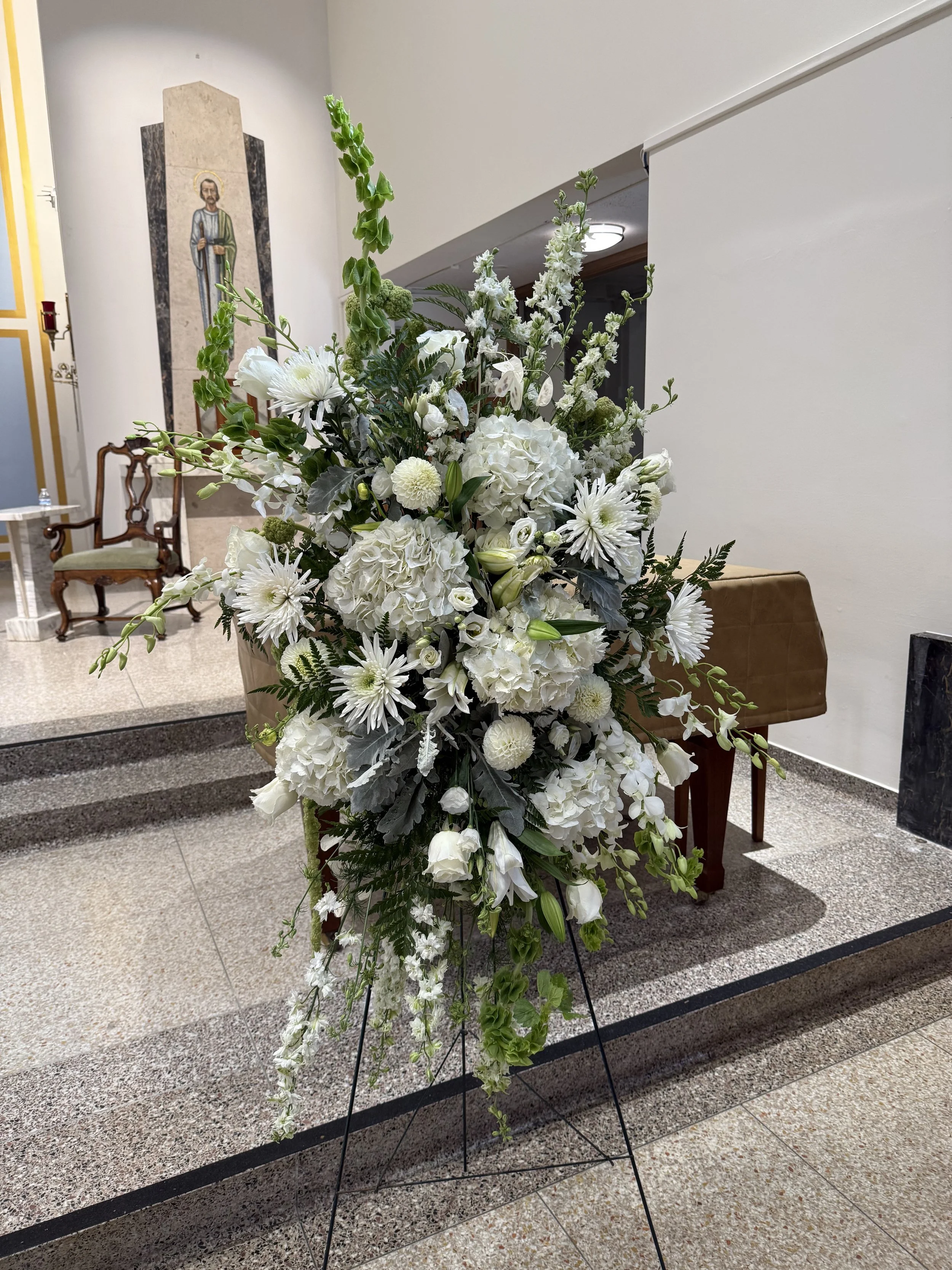 A large floral display with white flowers, including hydrangeas, lilies, roses, and daisies, arranged on a black metal stand in a room with a beige tiled floor and white walls.