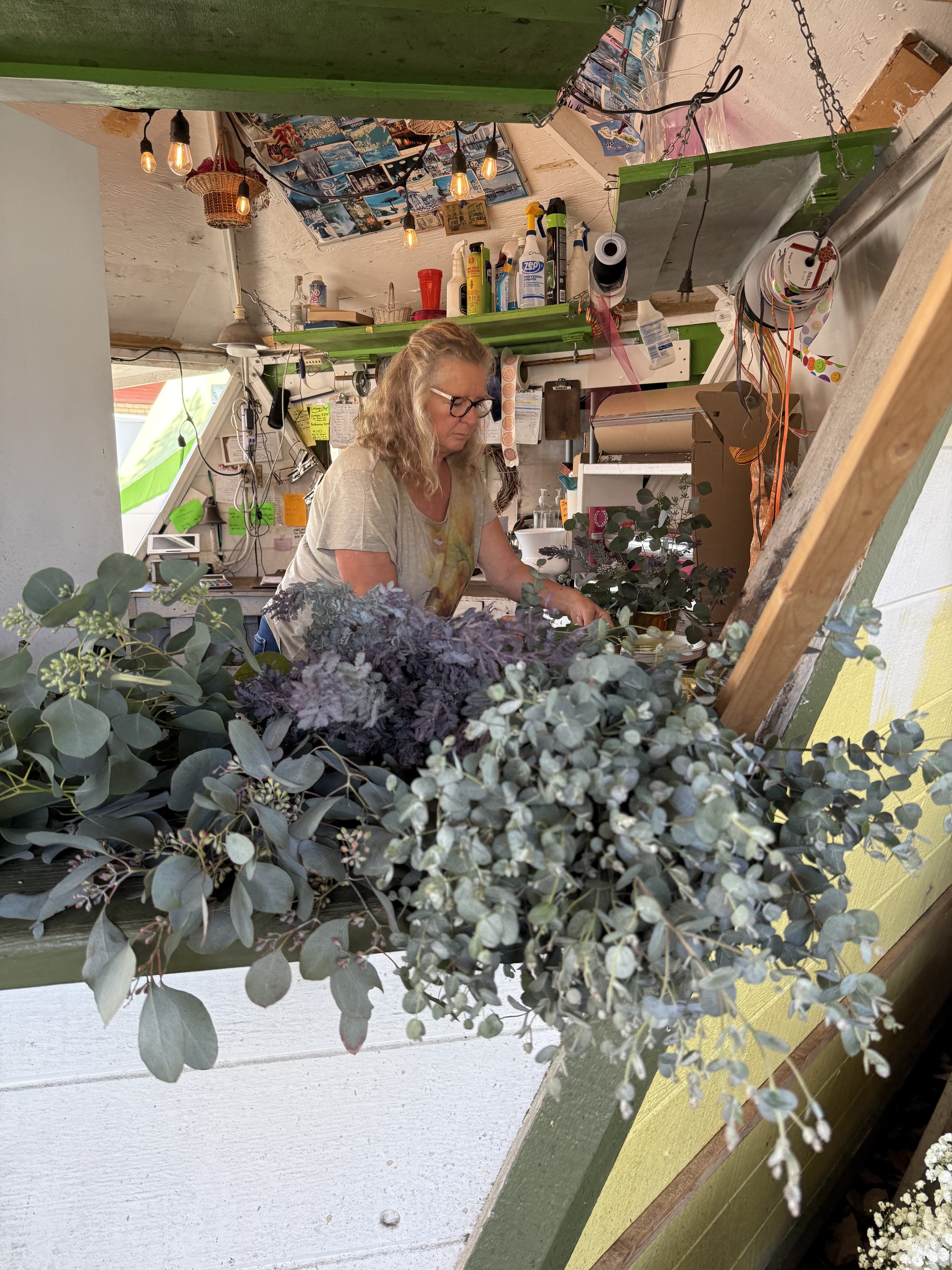 A woman with curly blonde hair and glasses working at a cluttered workbench in a small, brightly lit artist's or craftsman's studio. The space has colorful decorations, plants, and various tools and supplies on shelves.