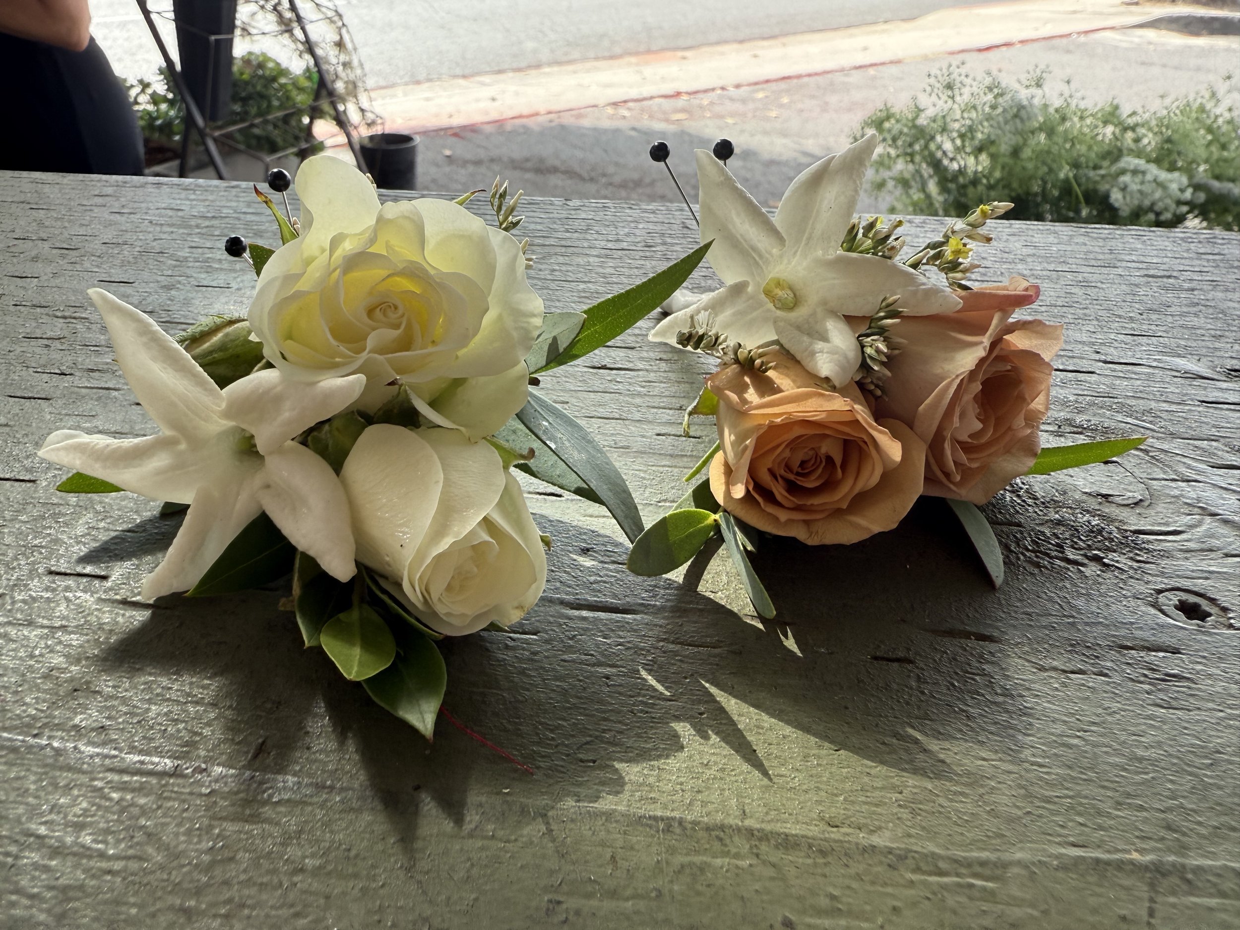 Two small floral arrangements with roses and white flowers on a rustic wooden table, outdoors with pavement and greenery in the background.