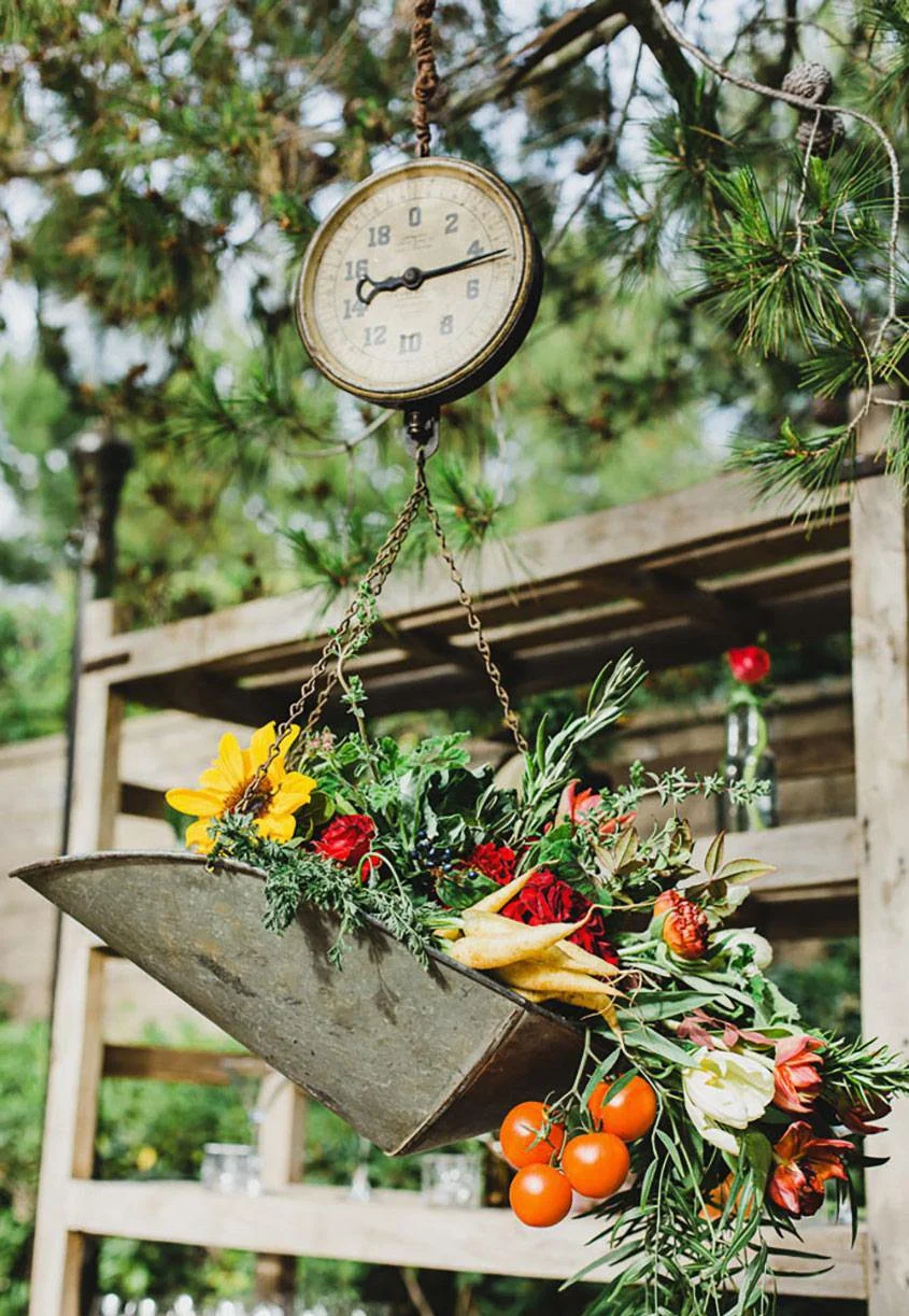 Hanging metal flower basket with assorted flowers and vegetables, under a clock in an outdoor garden setting.
