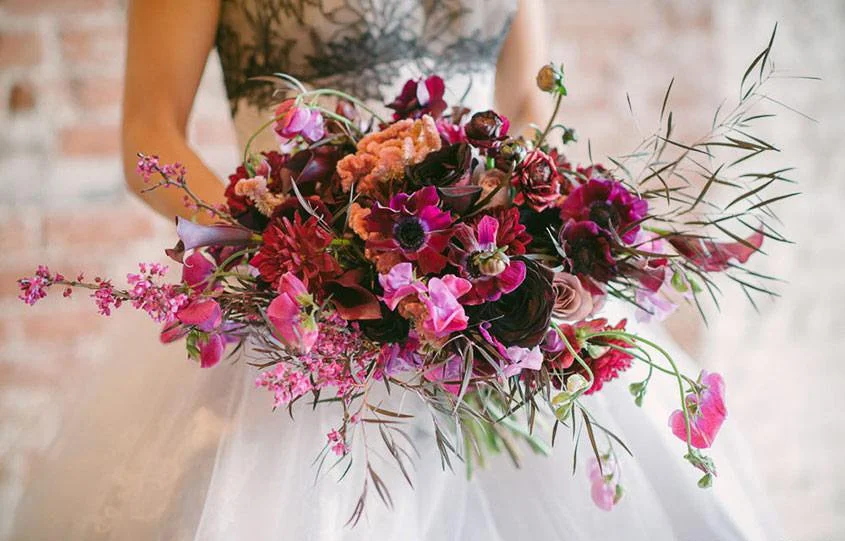 A person holding a large, colorful bouquet of pink, purple, and red flowers with green foliage, in front of a blurred indoor background.
