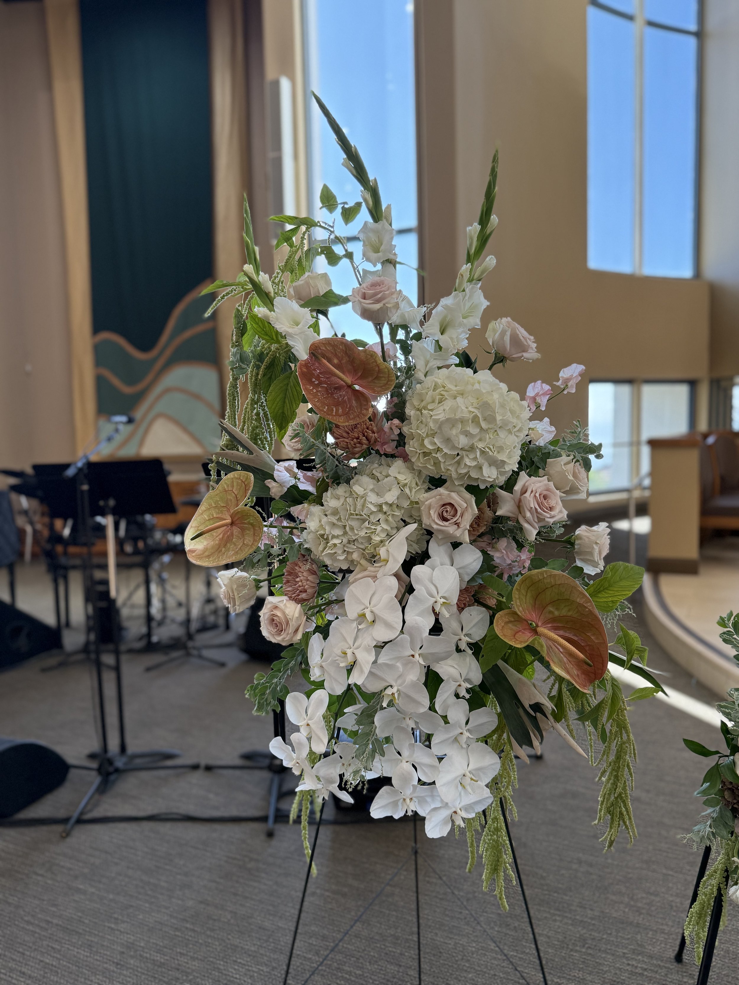 A large floral arrangement with white, pink, and reddish flowers on a stand in an indoor setting with windows and musical instruments in the background.