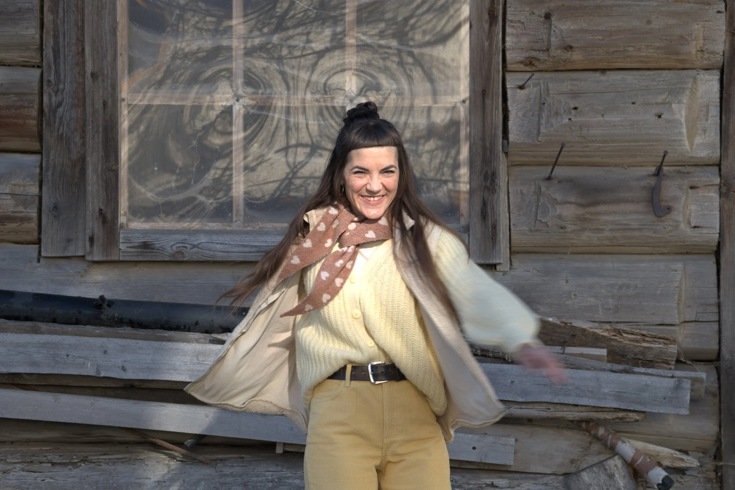 Junge Frau lächelt vor einem Holzhaus mit einem Fenster im Hintergrund.